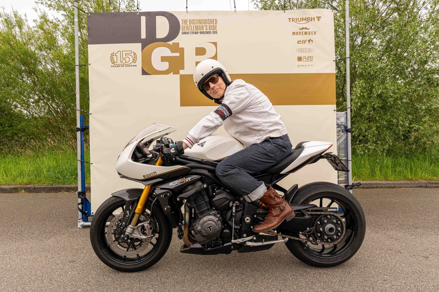 Rider in helmet and casual attire poses on a modern sport motorcycle at a racing circuit near a DGB banner.