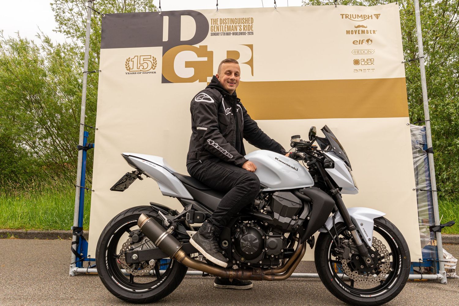 Man in black jacket posing on a silver sport motorcycle at a DGB motorsport event outdoors.