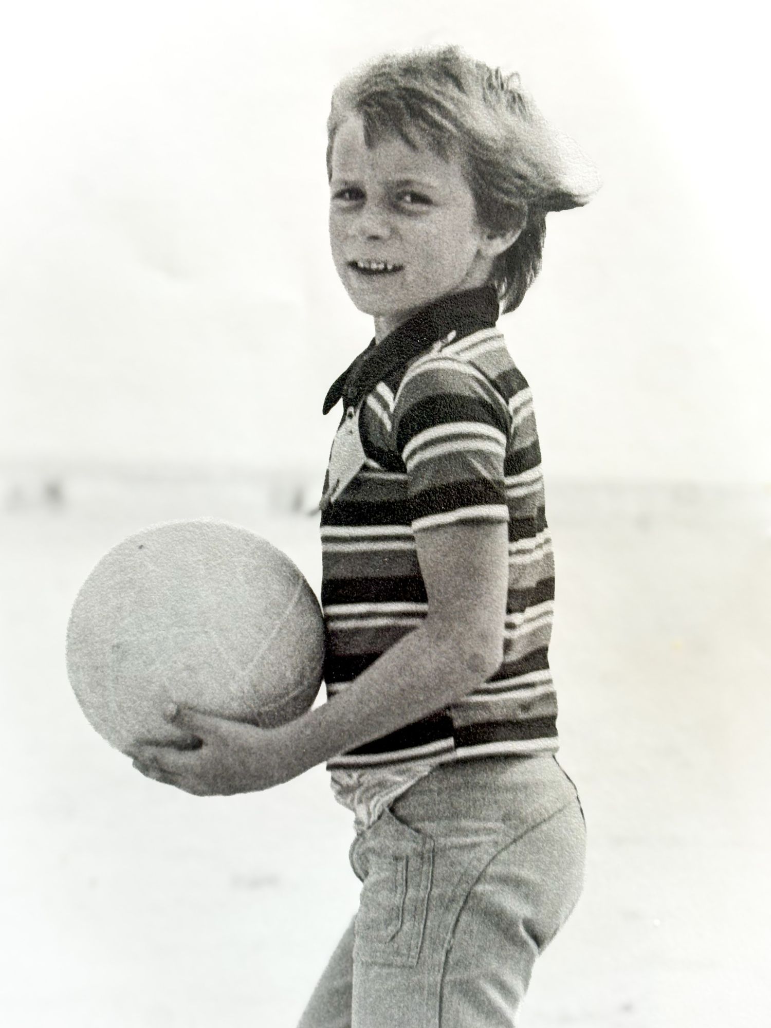 Young boy smiling on a beach holding a ball, wearing a striped polo shirt in a vintage black and white photo.