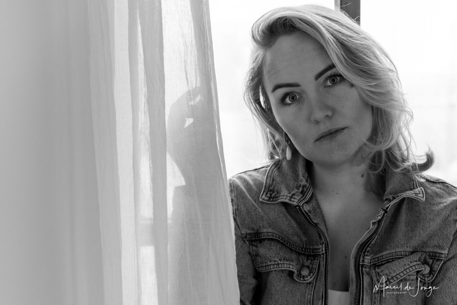 A black and white portrait of someone wearing a denim jacket while posing near a window with soft natural lighting.