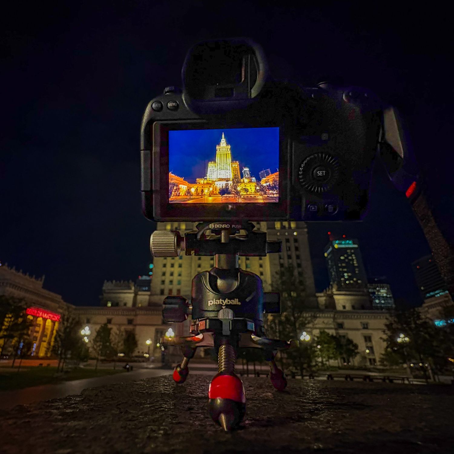 DSLR camera on tripod at night capturing illuminated skyscraper cityscape on LCD screen with dark blue sky backdrop.