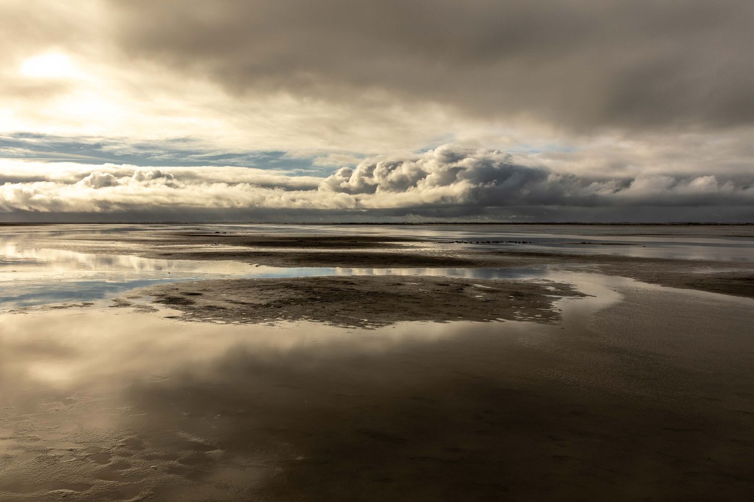 Dramatic storm clouds reflecting on wet sandy beach at low tide with golden light breaking through overcast sky.