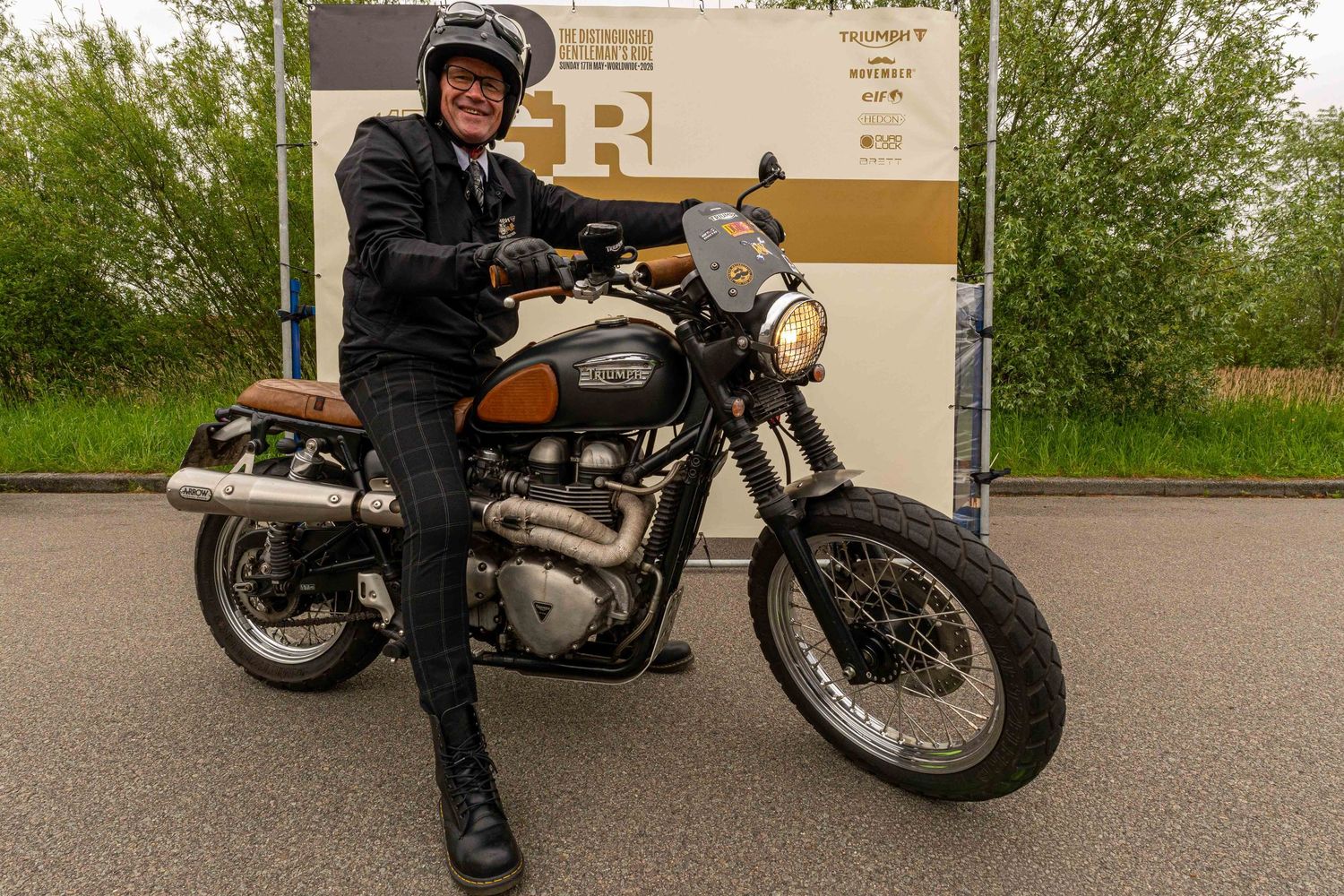 Person posing with a classic Triumph scrambler motorcycle outdoors near a branded banner.