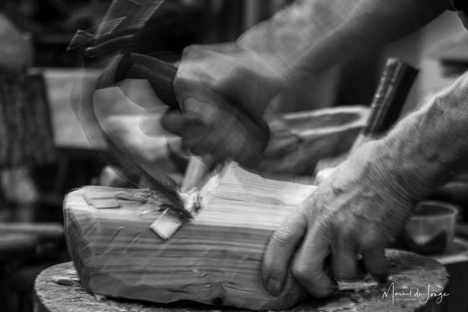 Hands carve wood with traditional tools in black and white artistic photograph showing woodworking craftsmanship.