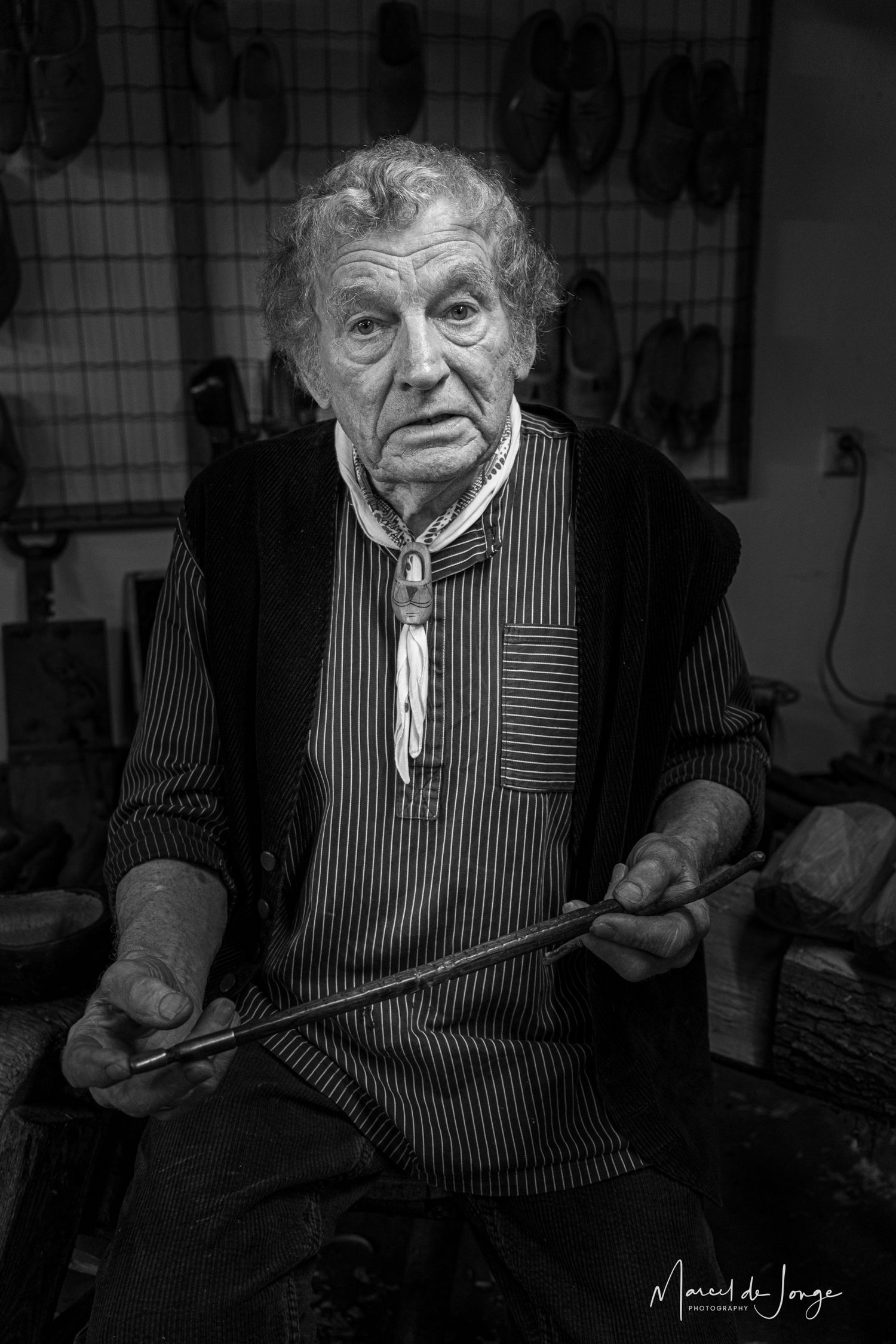 Black and white portrait of an elderly person in a striped shirt holding tools in a workshop setting.