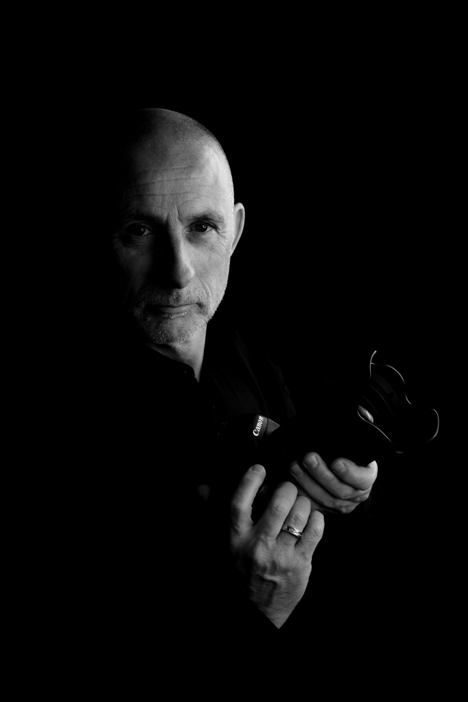 Dramatic black and white portrait of photographer in dark setting holding camera with strong contrast lighting.