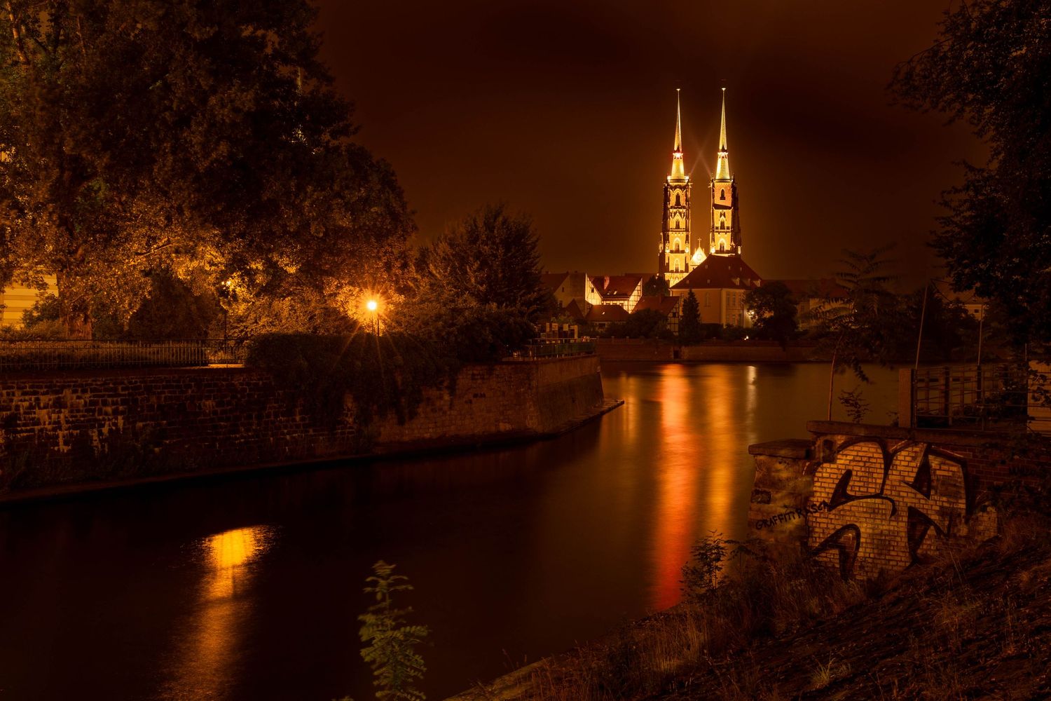 Illuminated Gothic cathedral spires reflect on a calm river at night, surrounded by glowing streetlights and lush trees.
