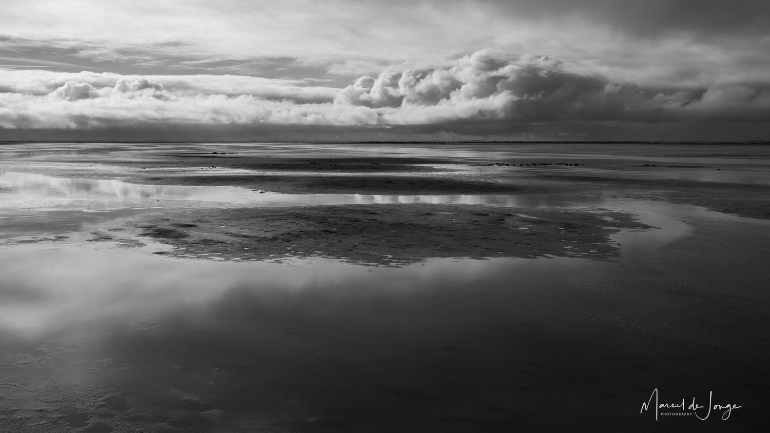 A dramatic black and white panoramic seascape shows rolling waves and moody clouds stretching across the horizon.
