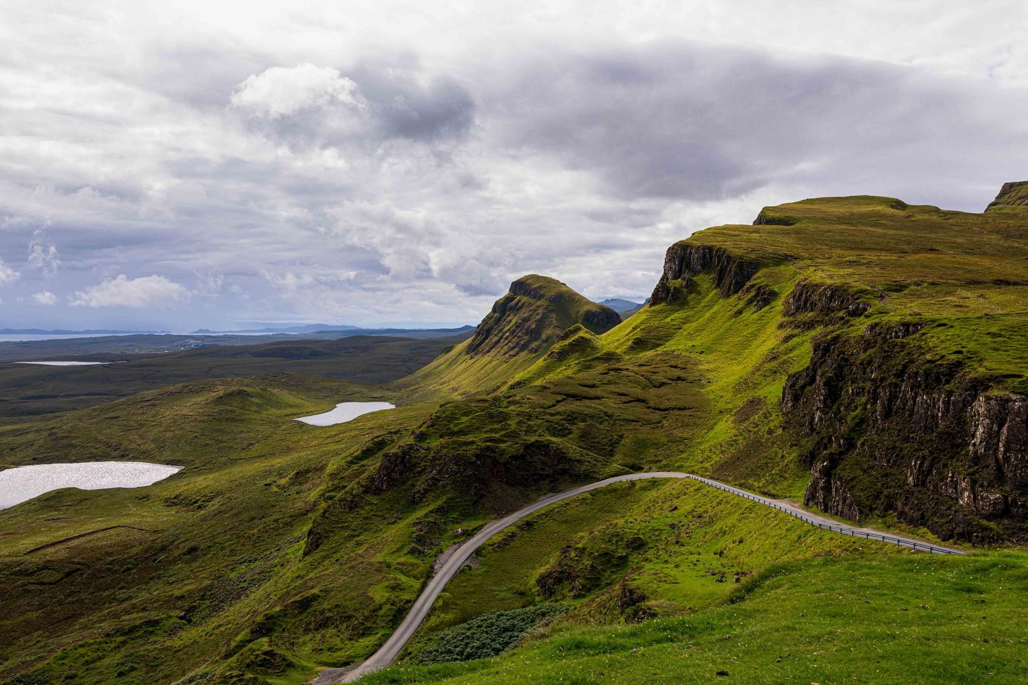 Aerial view of winding road through lush green Scottish Highlands with dramatic cloudy sky and serene lakes below.