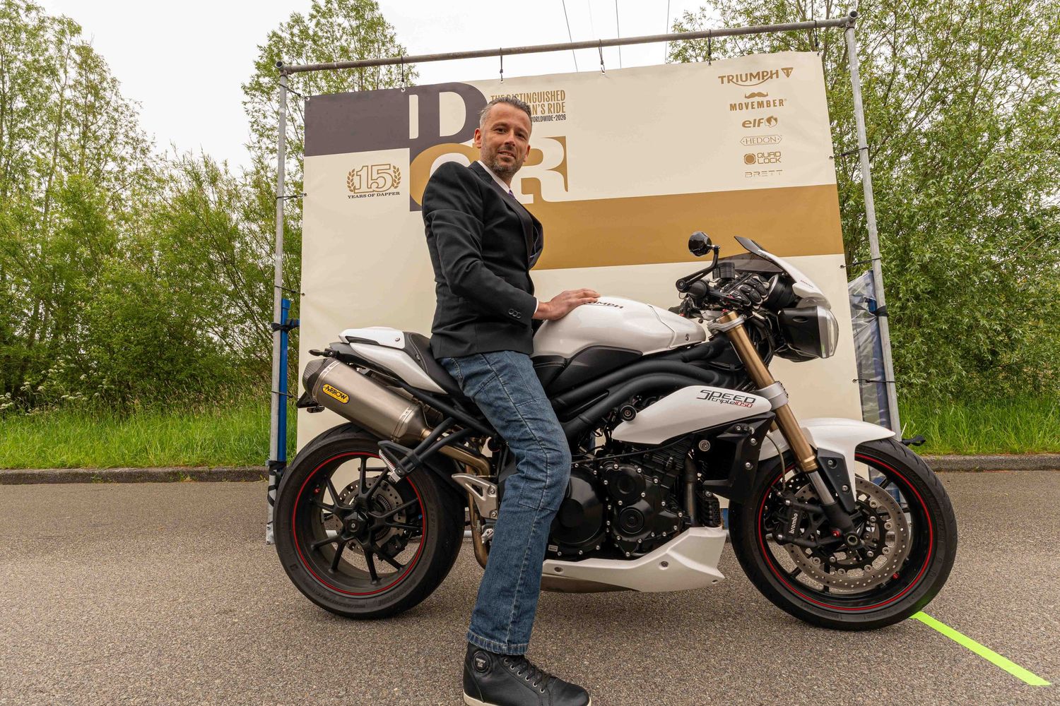 Man in black jacket stands beside a white Triumph Speed Triple motorcycle at an outdoor motorsport event.