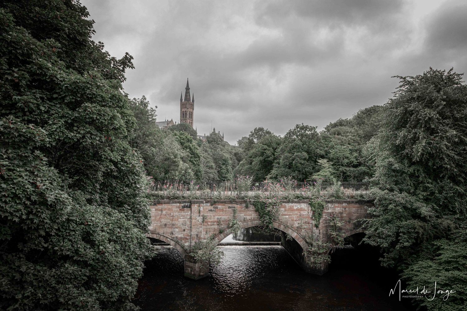 A panoramic view of a historic stone bridge and church spire under moody gray clouds surrounded by lush green trees.