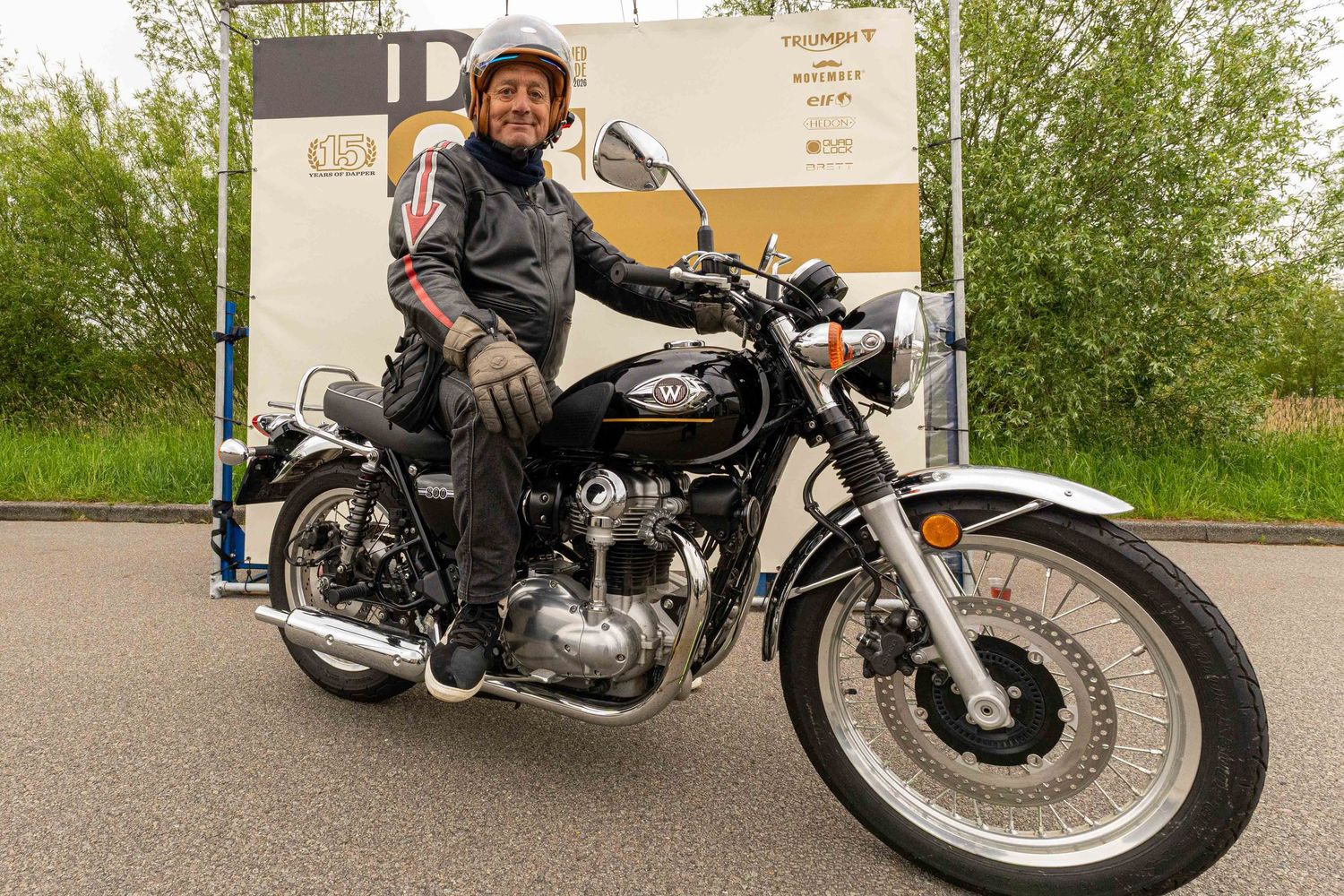 A smiling rider in a black leather jacket sits on a classic black motorcycle at an outdoor motorcycle event.