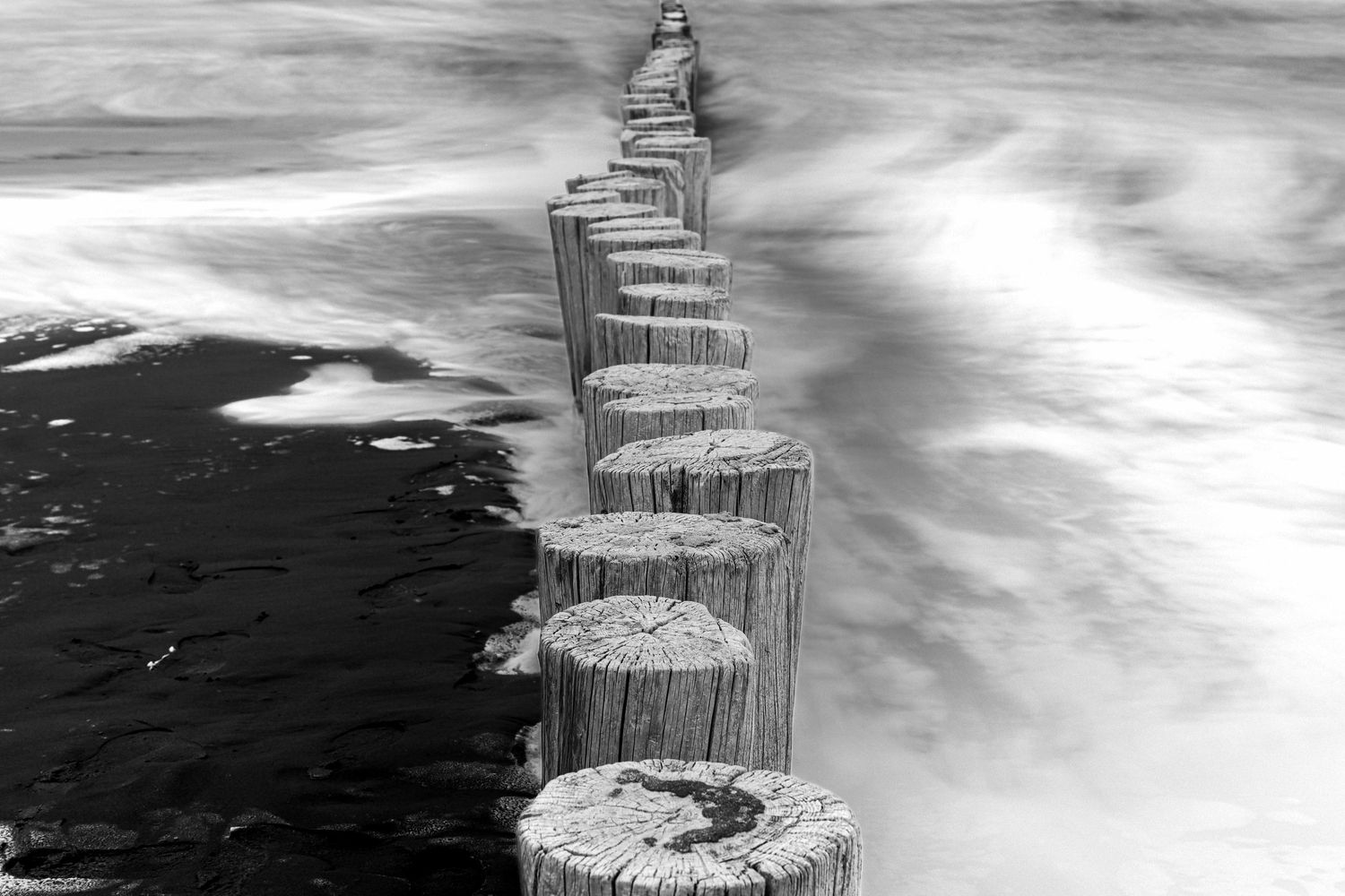 Black and white photo of weathered wooden posts extending into misty ocean waves, creating a serene coastal scene.