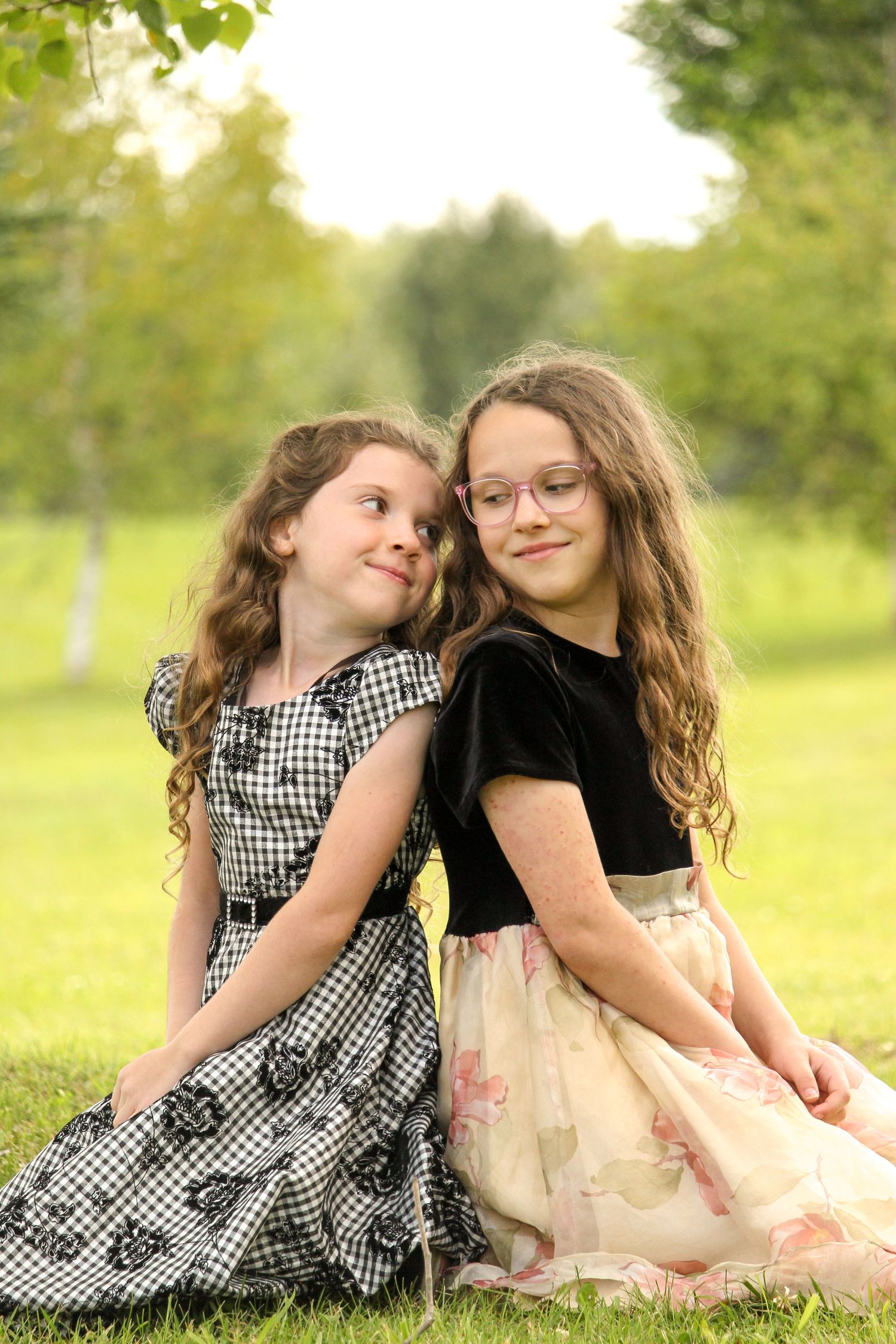 Two young girls sitting in grass back-to-back looking at each other, Rocky Mountain House, AB