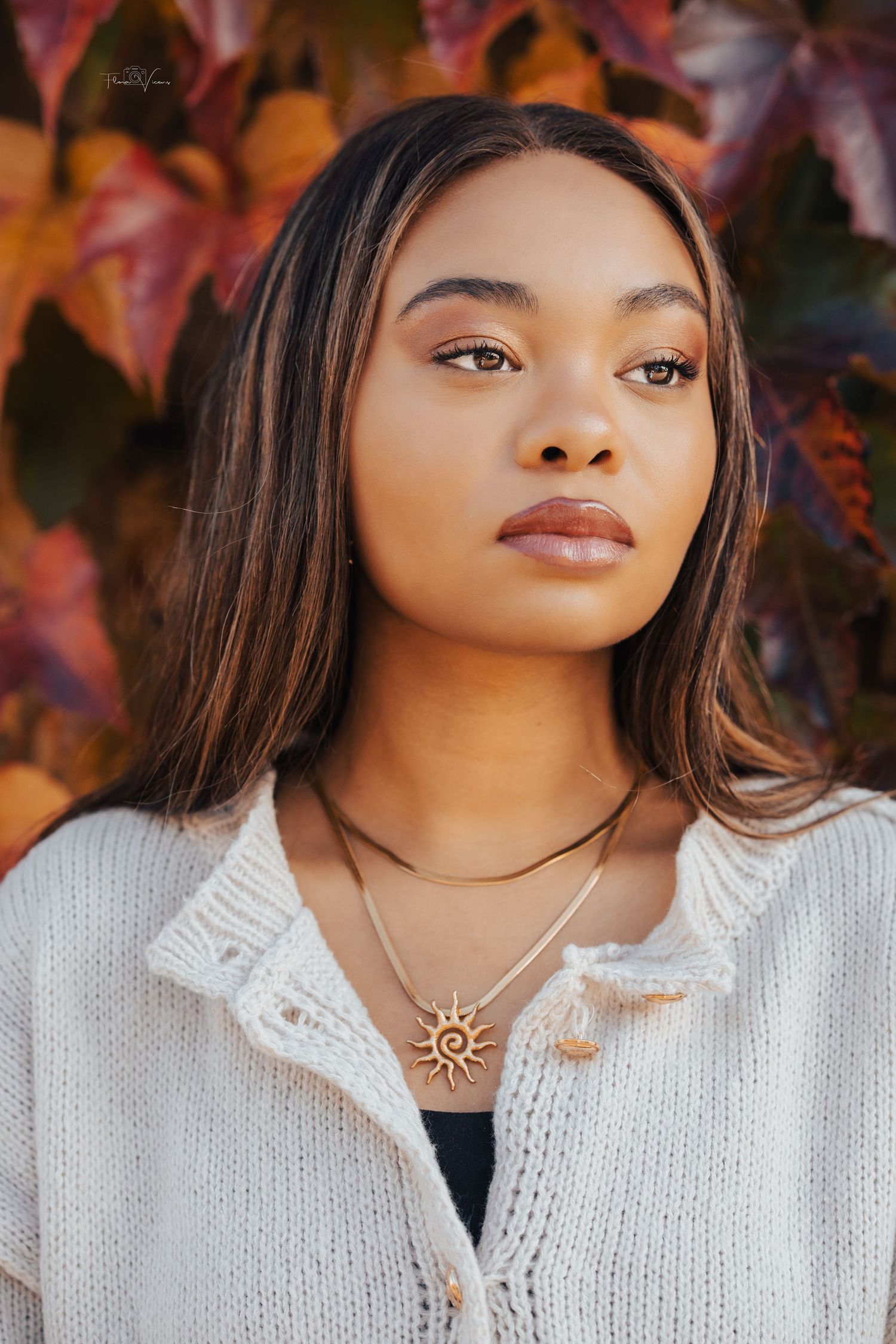 Portrait against autumn foliage wearing a white knit sweater and sun pendant necklace.
