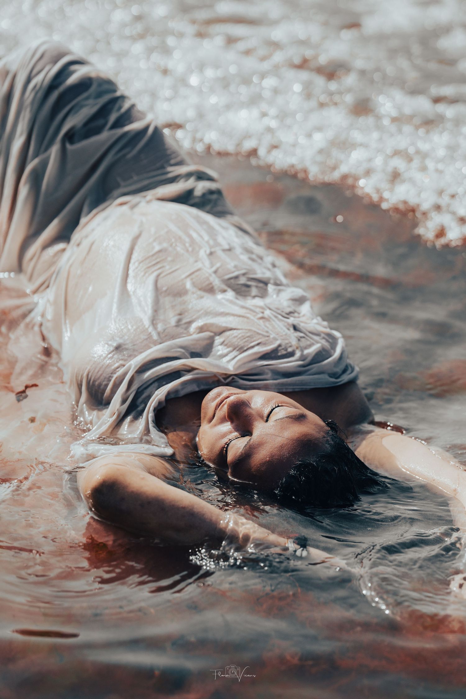 Woman in white dress lying peacefully in shallow water with gentle waves, eyes closed, smiling serenely.