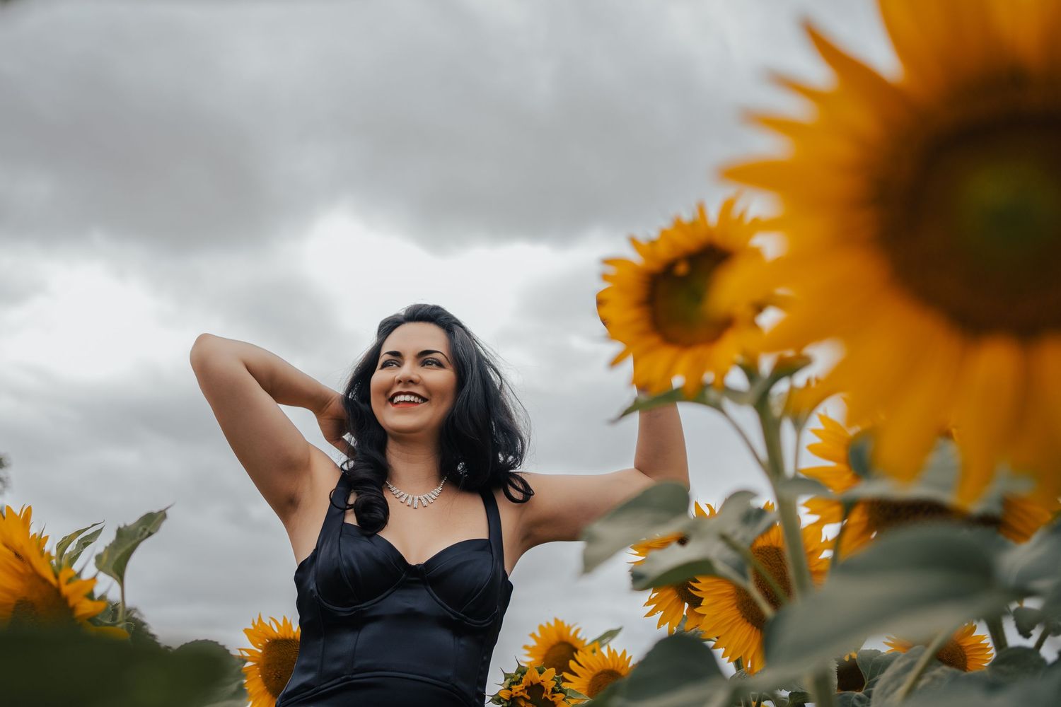 Woman in black top poses joyfully in blooming sunflower field under cloudy sky.