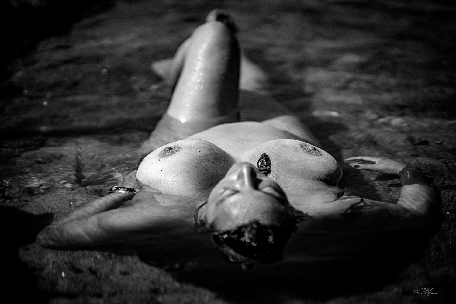 Black and white fine art nude photograph of a woman reclining on wet rocks outdoors.