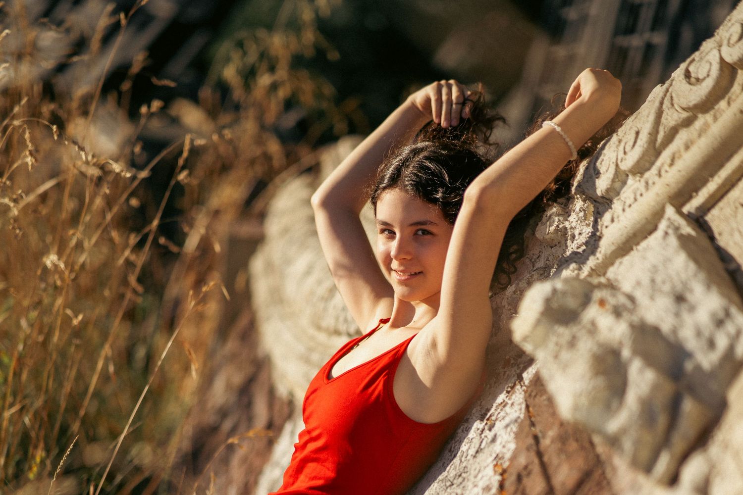 Woman in red dress leans against rocky wall in moody outdoor photoshoot.