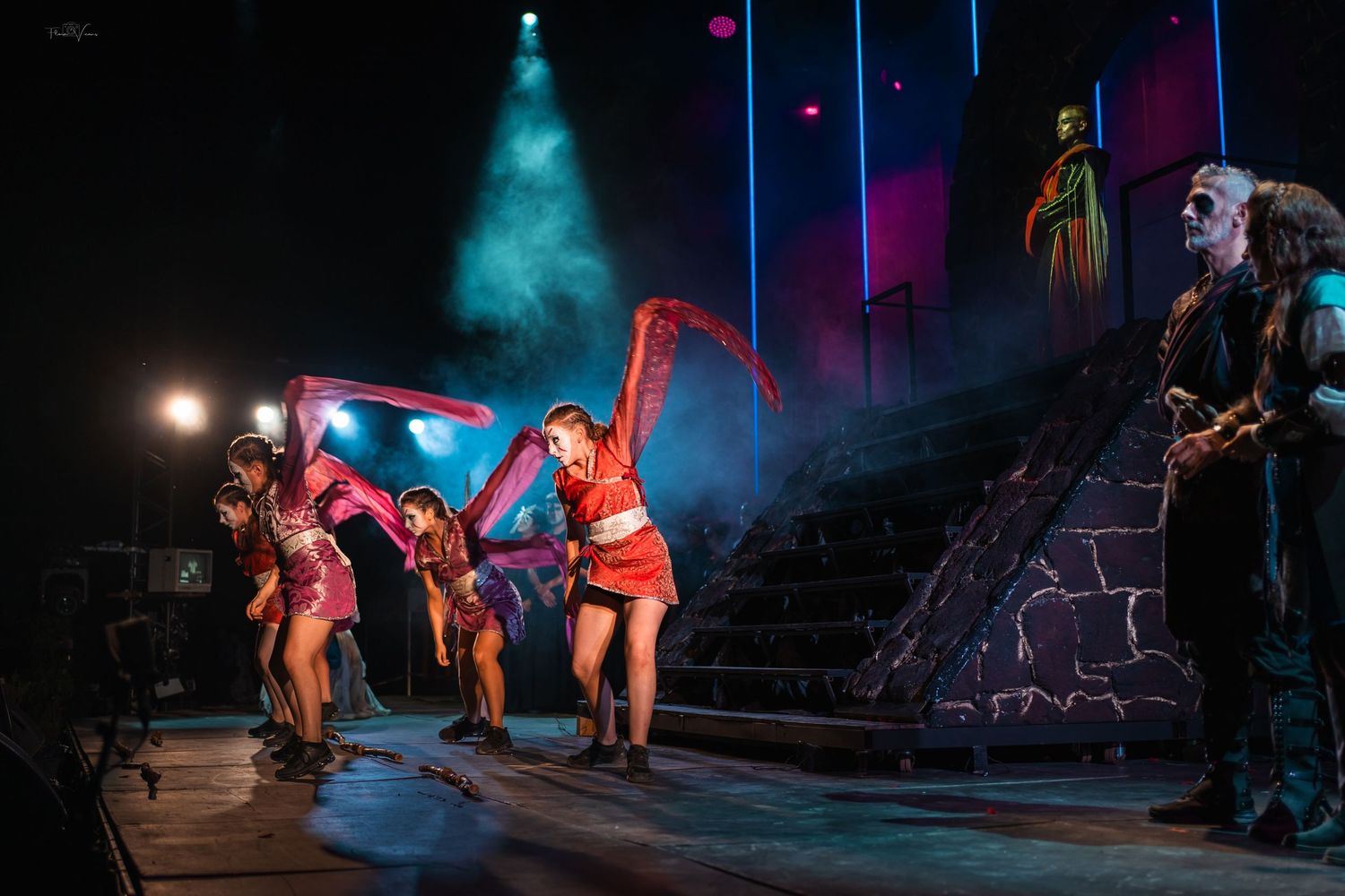 Performers dance energetically on a dimly lit stage with dramatic blue and red lighting effects.