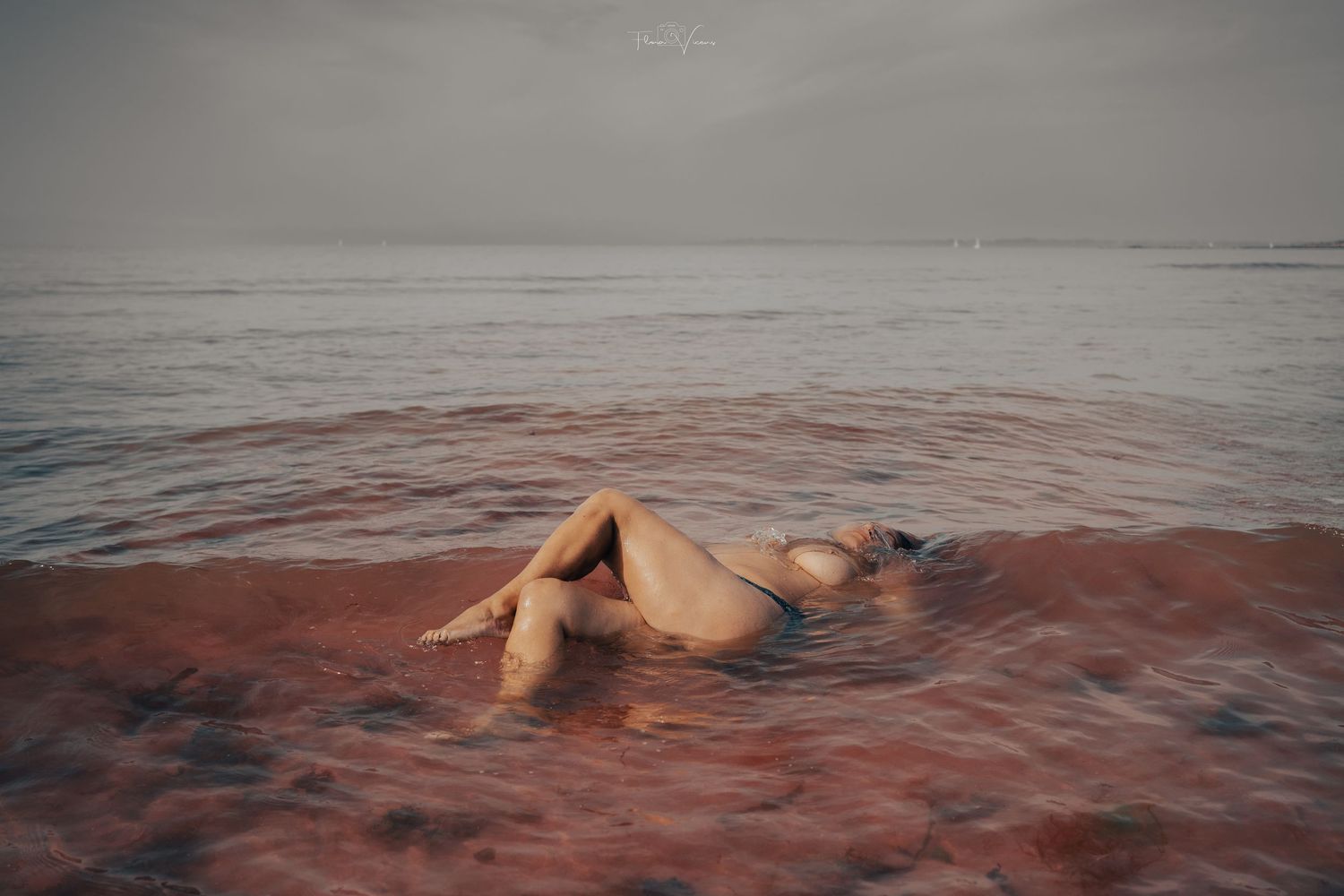 A nude figure reclines in shallow ocean waves on a sandy shore under an overcast sky.