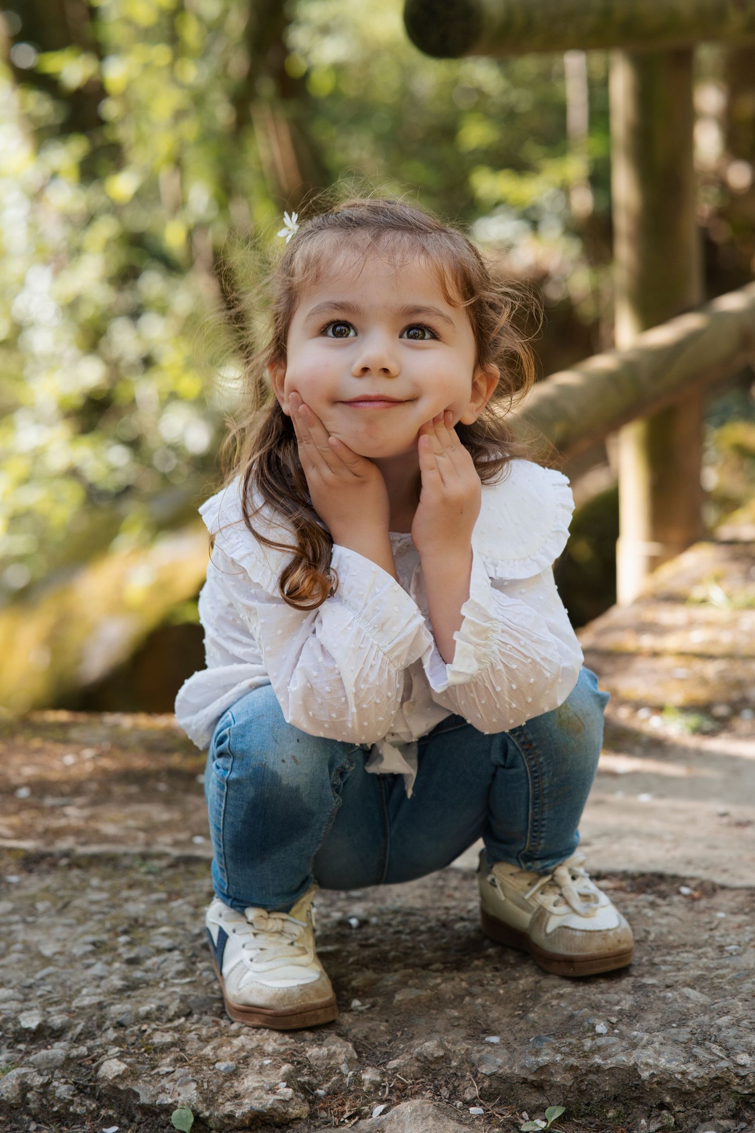 Child in white lace and denim crouching on a rock in a forest setting.