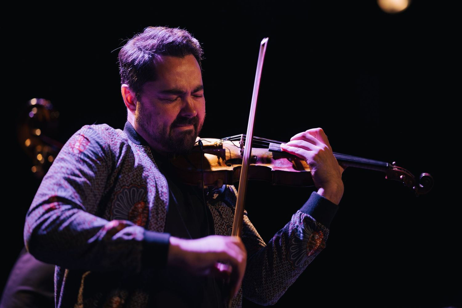 Musician performing with a string instrument against a dark background with dramatic lighting.