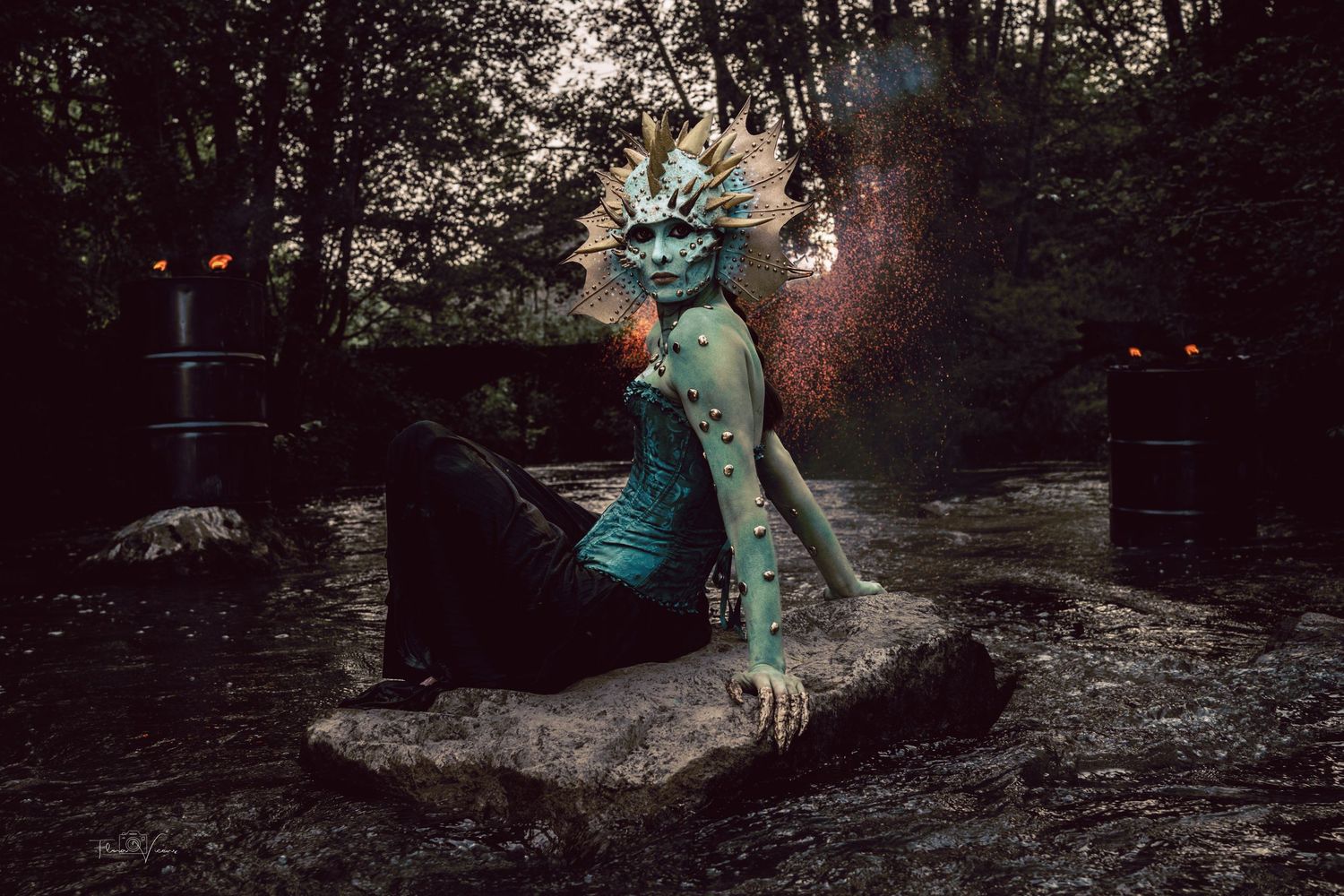 Fantasy creature costume with teal body paint and spiky headdress posed on a rock in a dark forest setting.