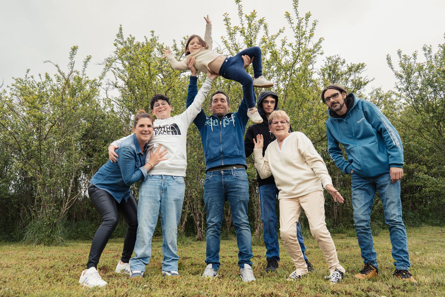 A playful group poses lying down in grass wearing casual denim and white outfits.