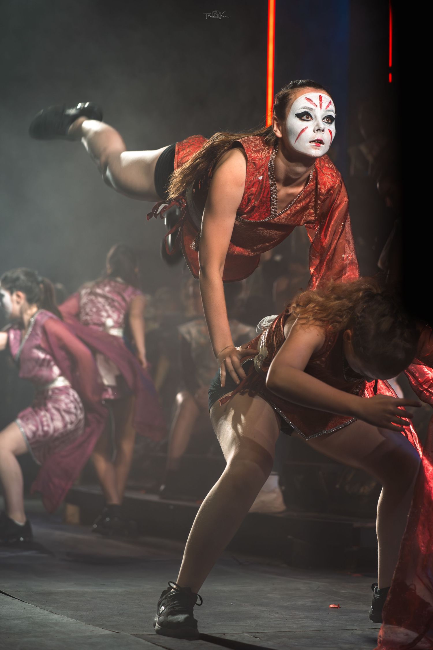 Dancers in red costumes perform a dynamic choreographed sequence on a smoky stage.