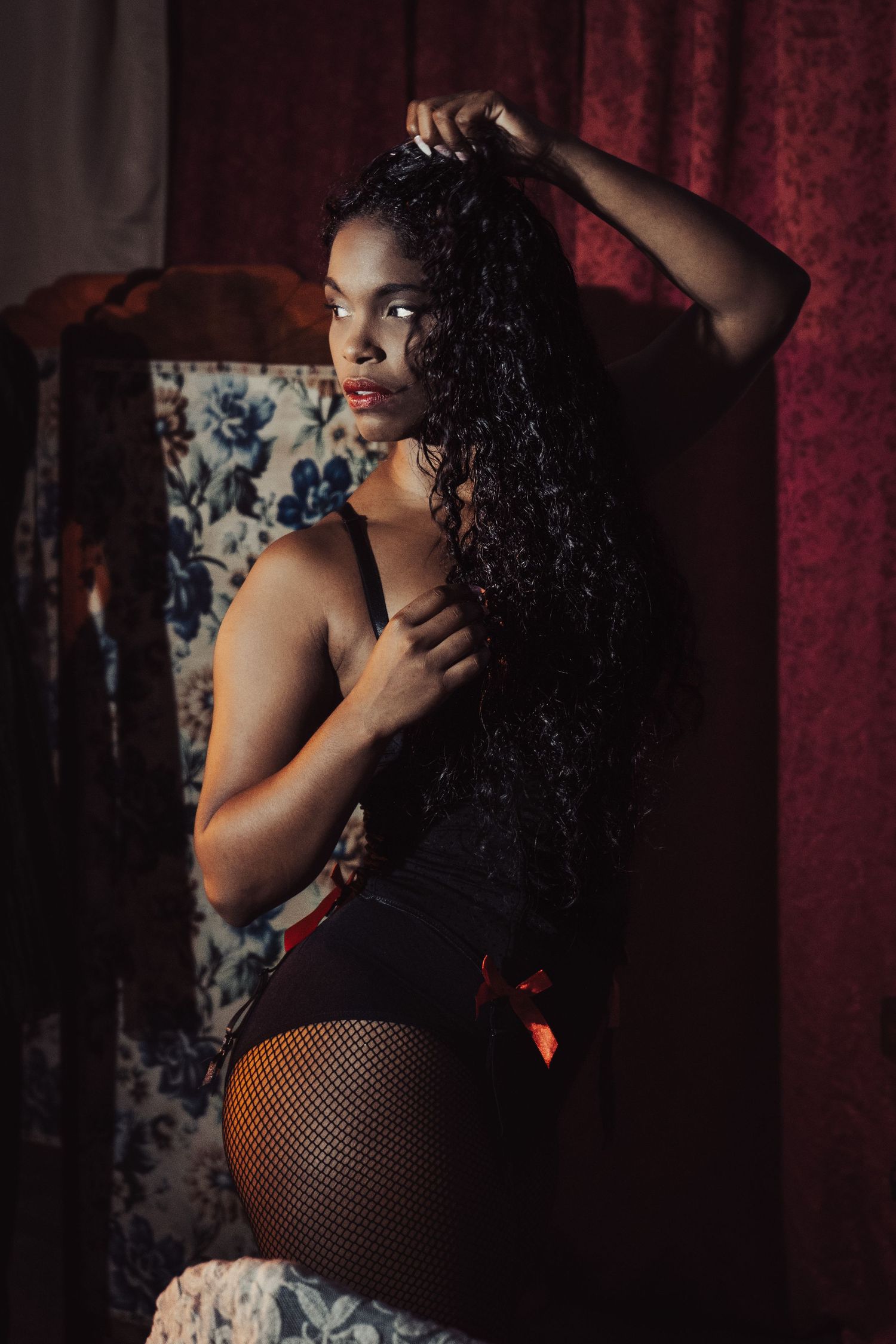 Moody boudoir photoshoot in dimly lit room with red curtains and patterned furniture.