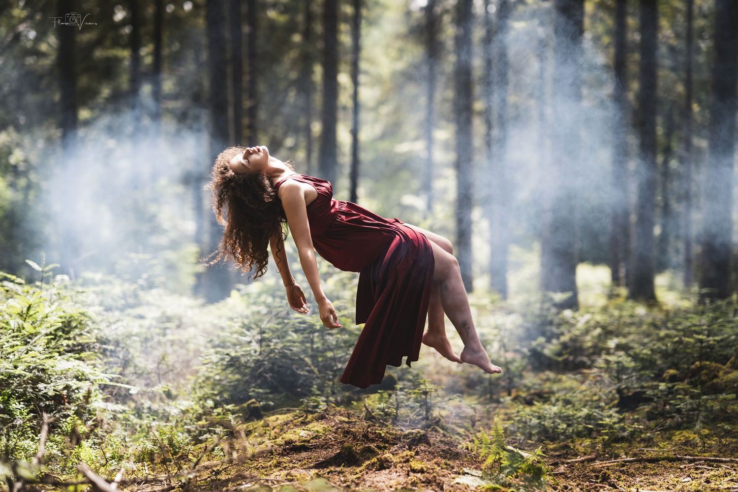 Woman in burgundy dress levitating in a misty sunlit forest with rays of light filtering through trees.