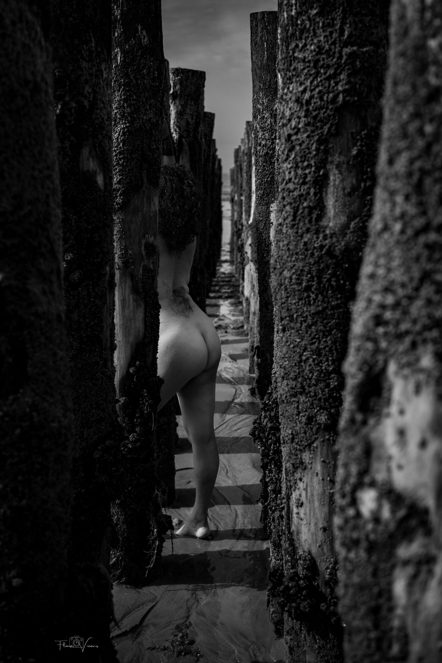 Black and white fine art nude photography of a figure walking between ancient mossy stone ruins toward light.