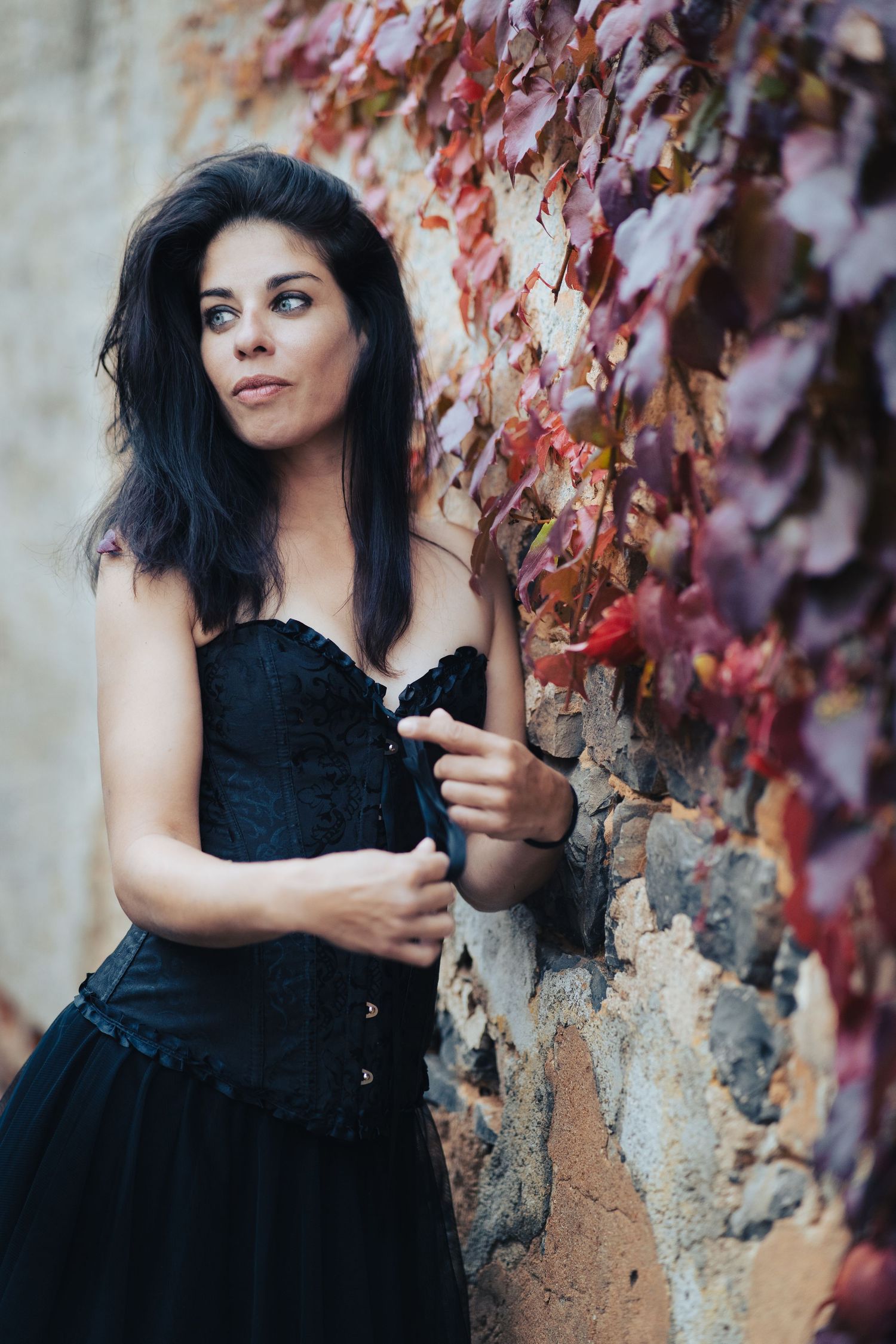 Dark-haired figure in black dress poses against weathered wall with red autumn vines.