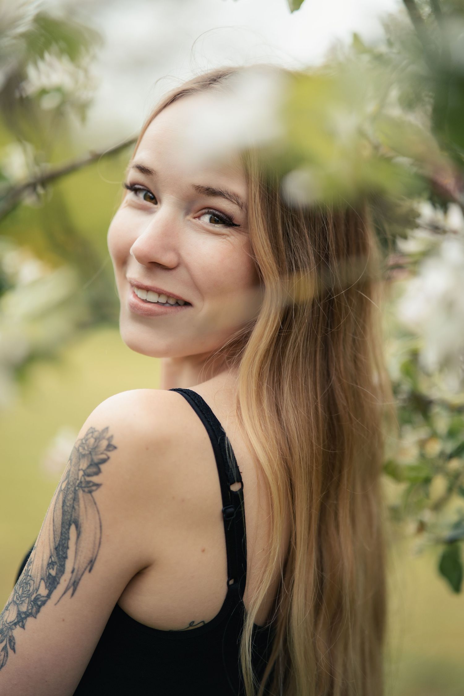A person with long blonde hair and shoulder tattoos poses outdoors in natural lighting.