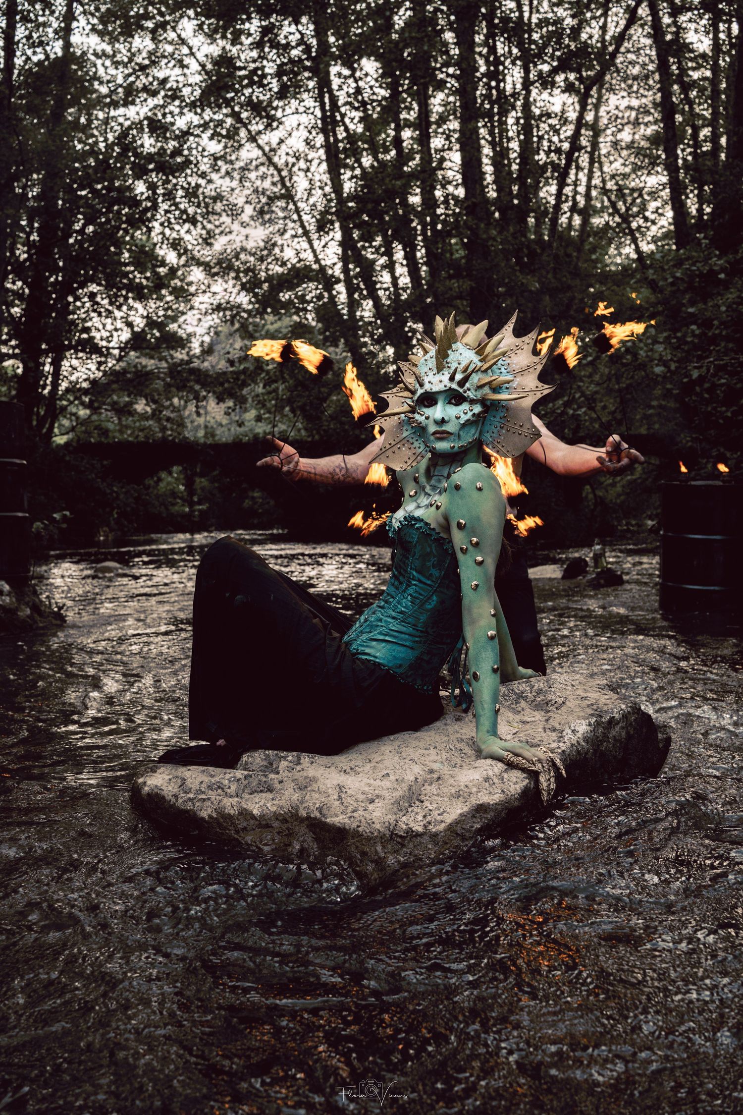 Fantasy creature cosplay with teal body paint, sun headdress, and fire props posed on a rock in a forest stream.