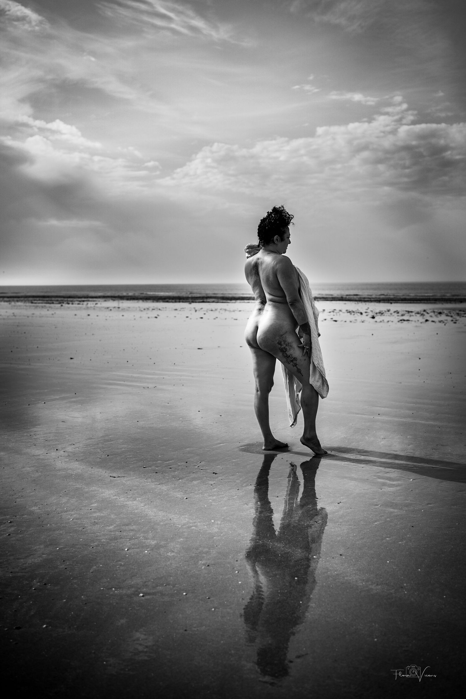 Black and white photo of a nude person standing on a reflective beach, holding fabric, facing the ocean under a cloudy sky.