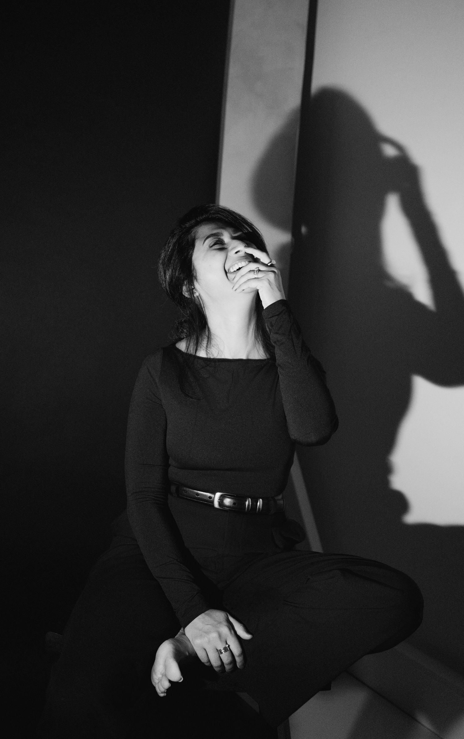 Black and white portrait of a laughing woman in black outfit with belt, sitting against wall with dramatic shadow.