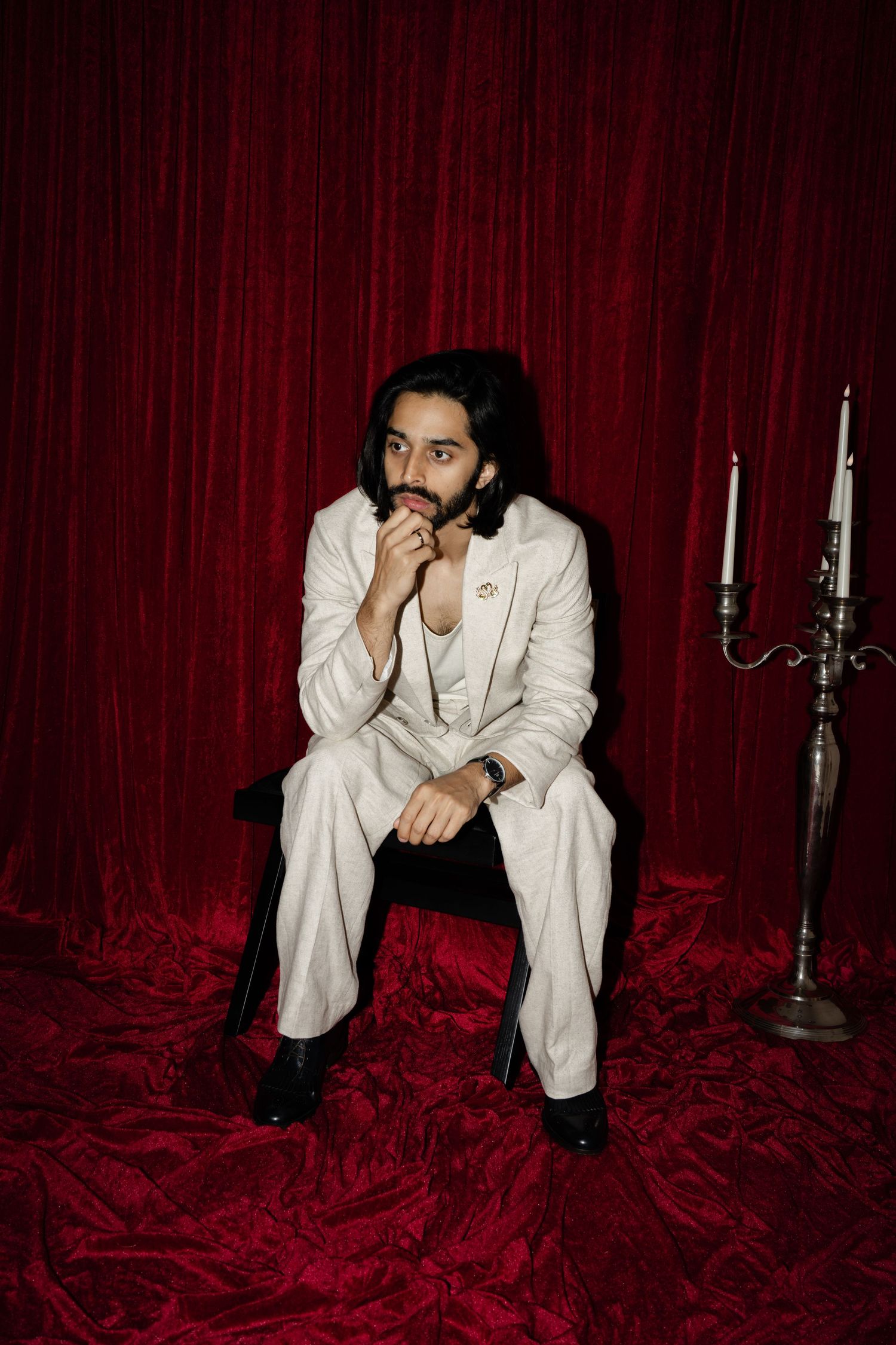 A figure in a white suit poses dramatically against red velvet curtains with silver candlesticks in a moody photo series.
