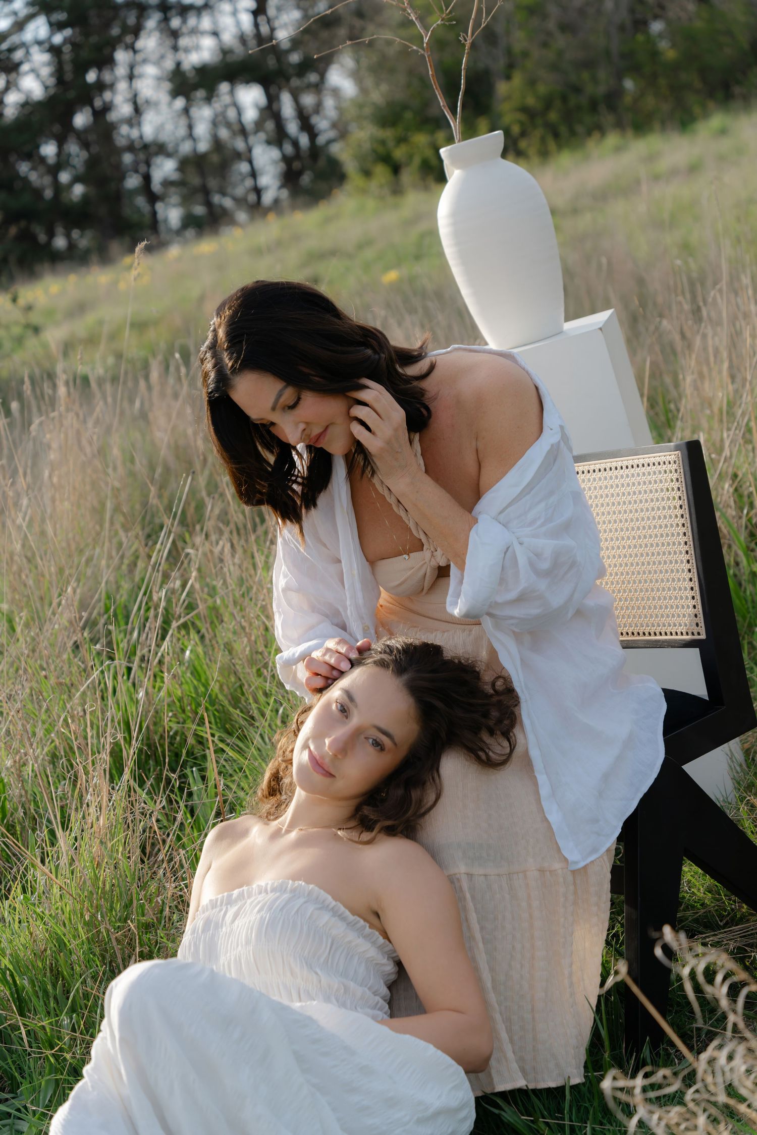 Two people in white clothing posed with decorative vase on chair in outdoor meadow setting.