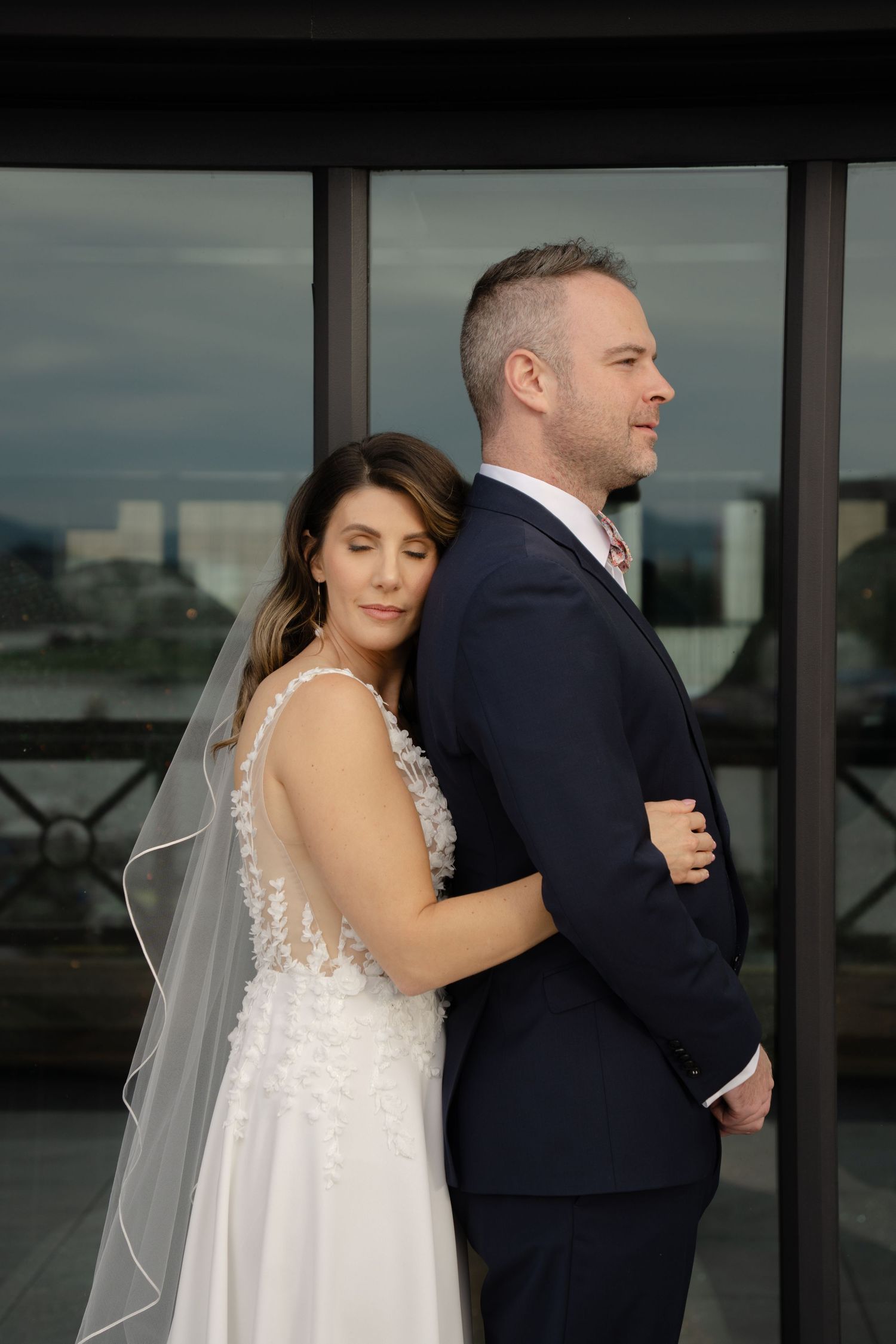 Elegantly dressed couple shares an intimate first look moment with city backdrop through large windows.