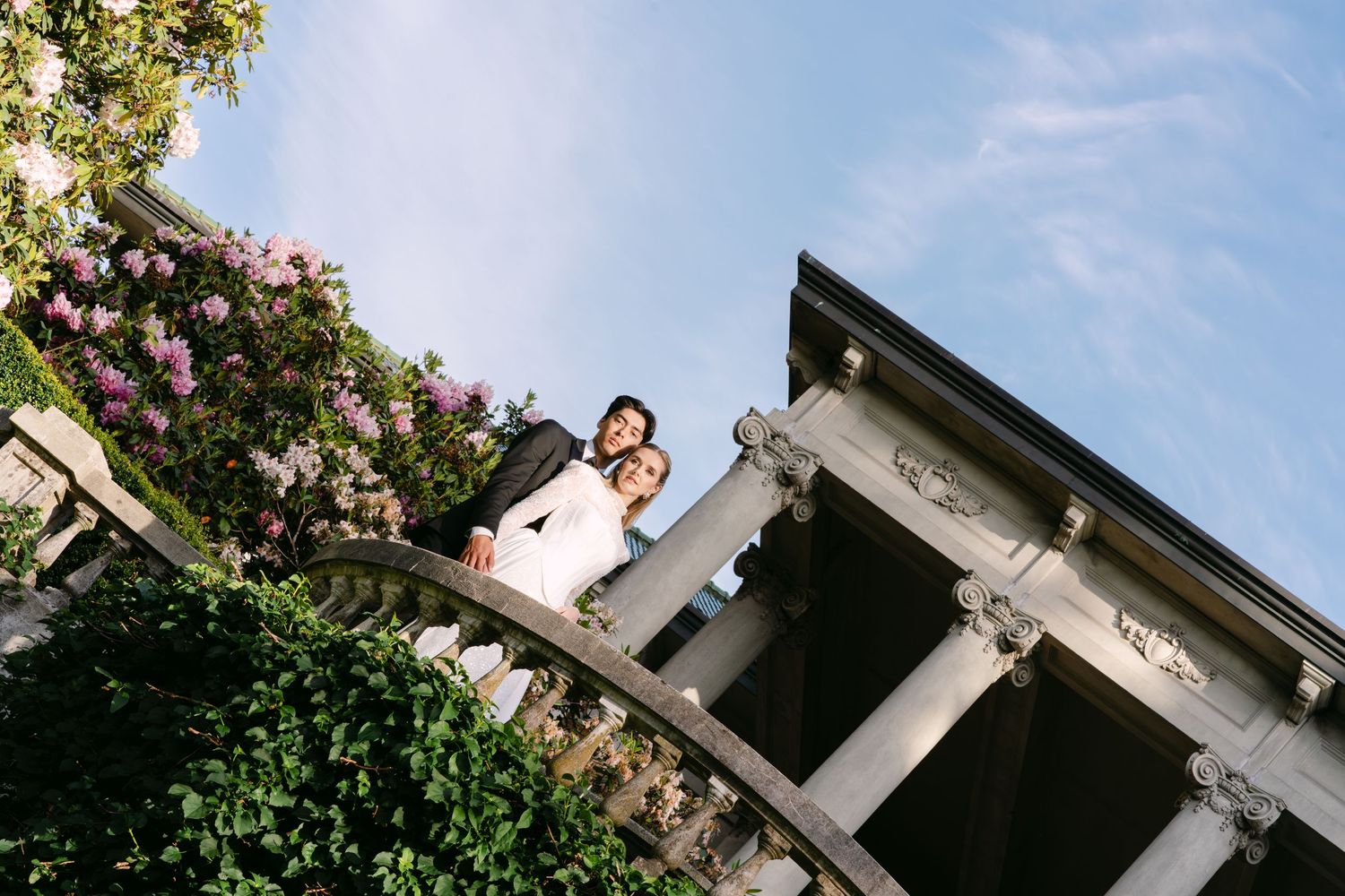 Wedding couple embraces on ornate balcony surrounded by blooming flowers and lush greenery under blue sky.