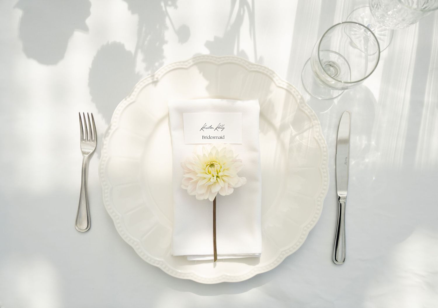 Minimalist place setting with white plate, silver cutlery and single yellow flower on white tablecloth.