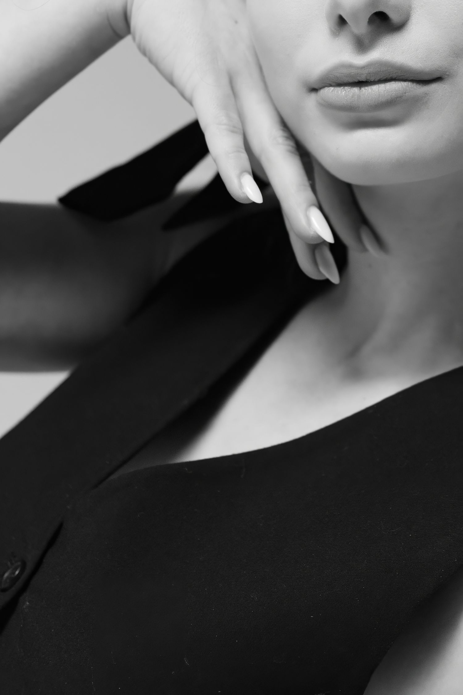 Black and white close-up of a woman in a black dress, hand near chin, with manicured nails and subtle lips.