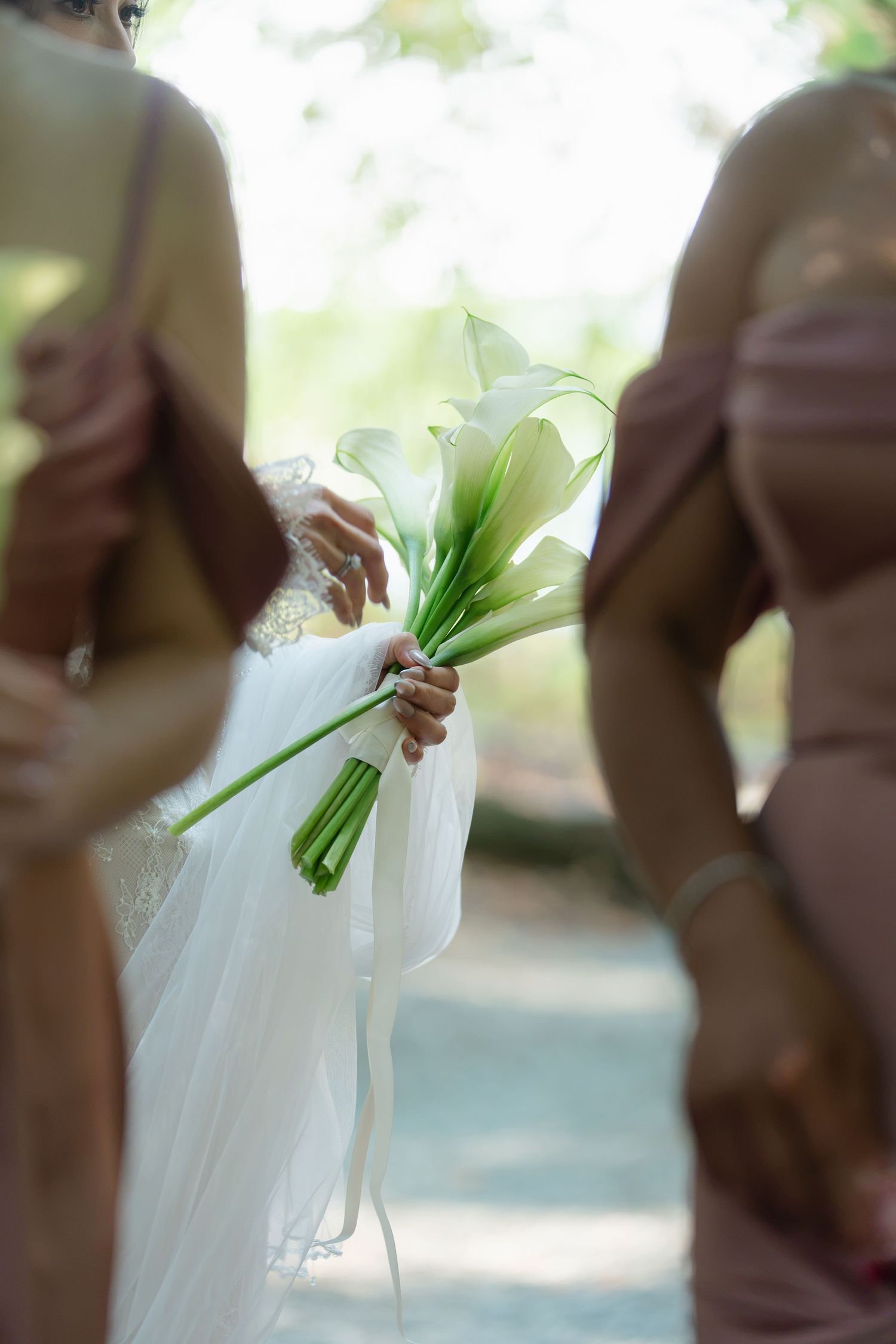Bride holding white calla lily bouquet with ribbons, flanked by bridesmaids in pink dresses at outdoor wedding.