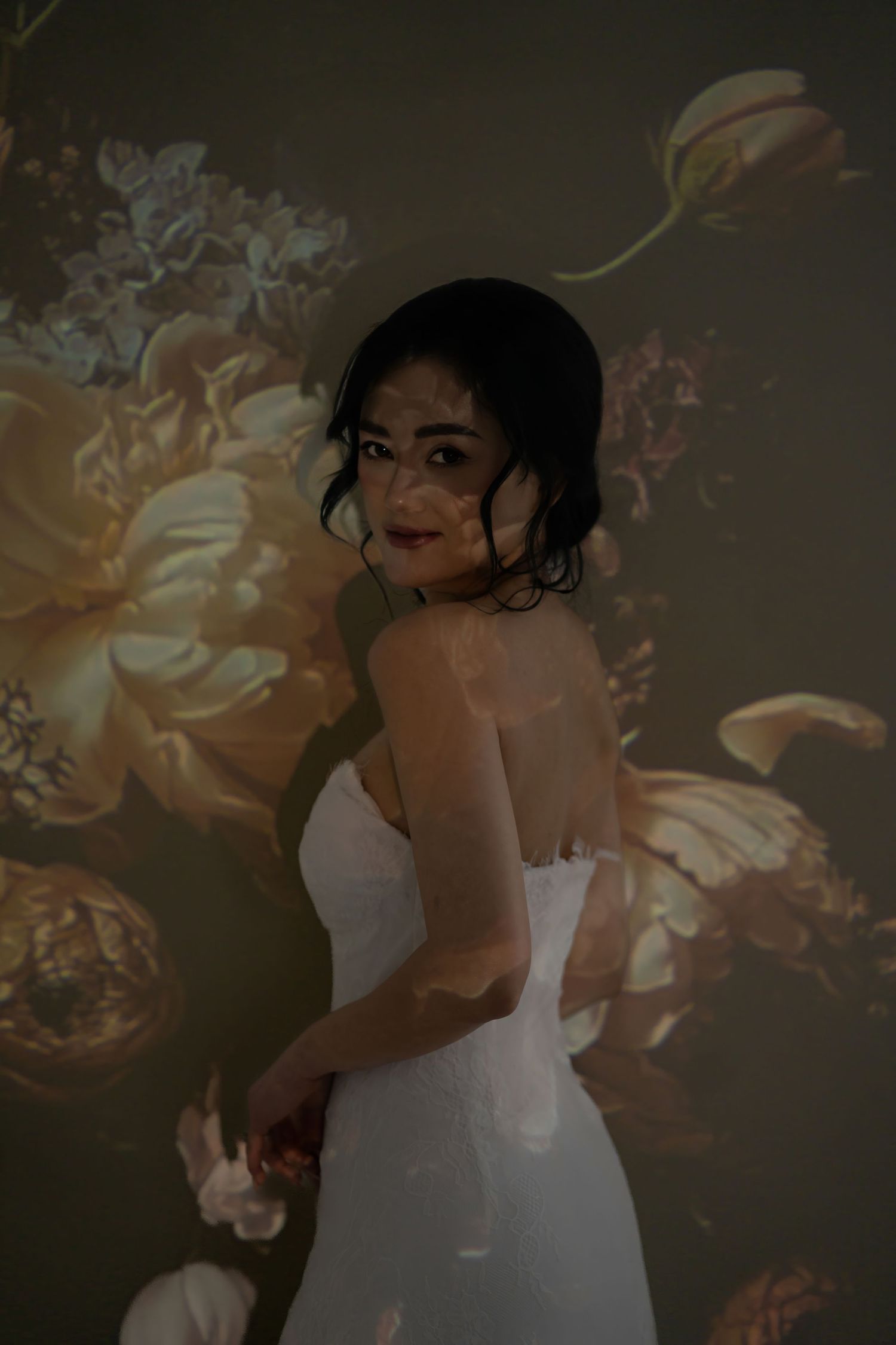 A bride in a white strapless gown poses against a floral projection backdrop in a moody, artistic portrait.