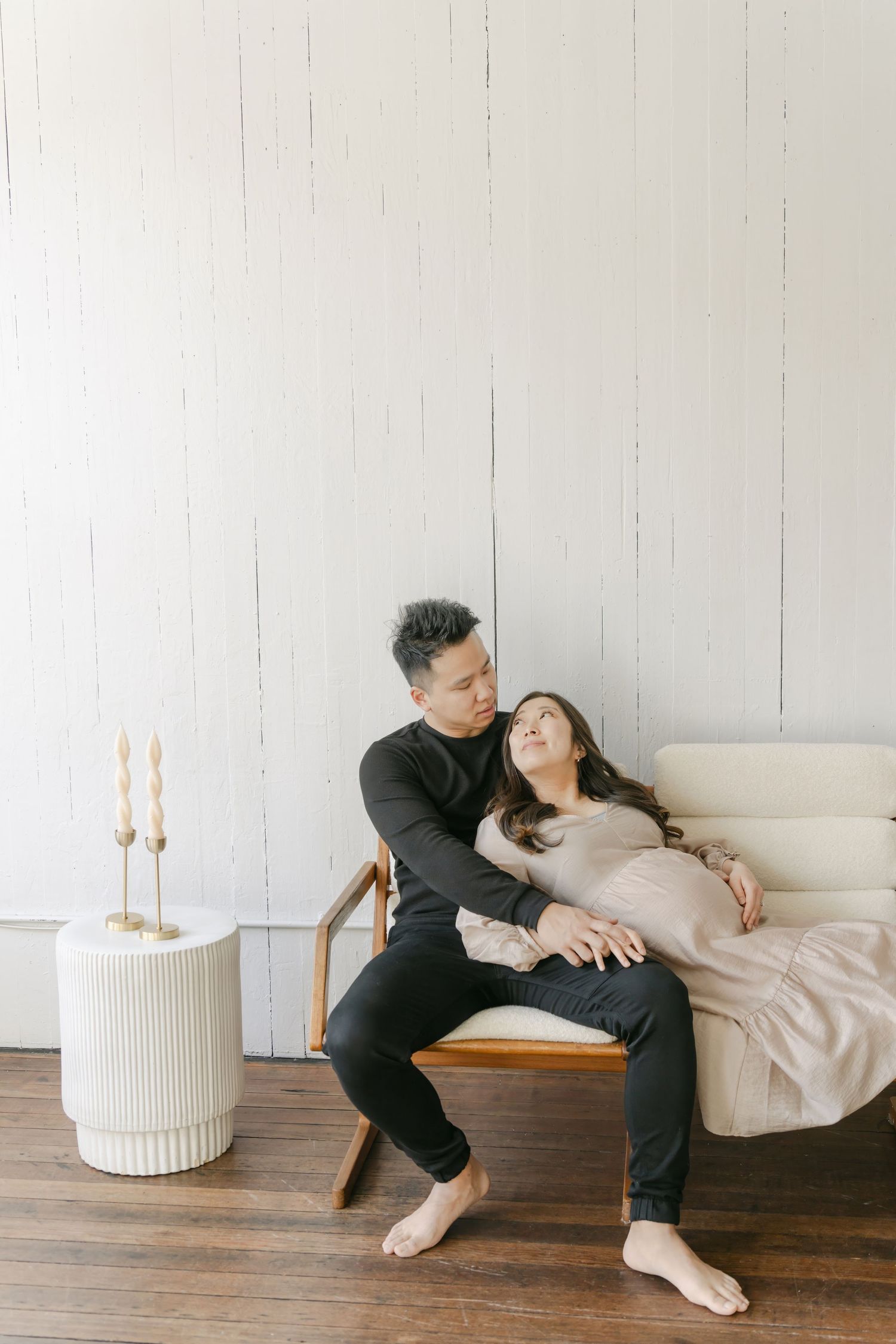 Couple in black clothing lounging together on a beige couch in a minimalist white room with candlesticks.