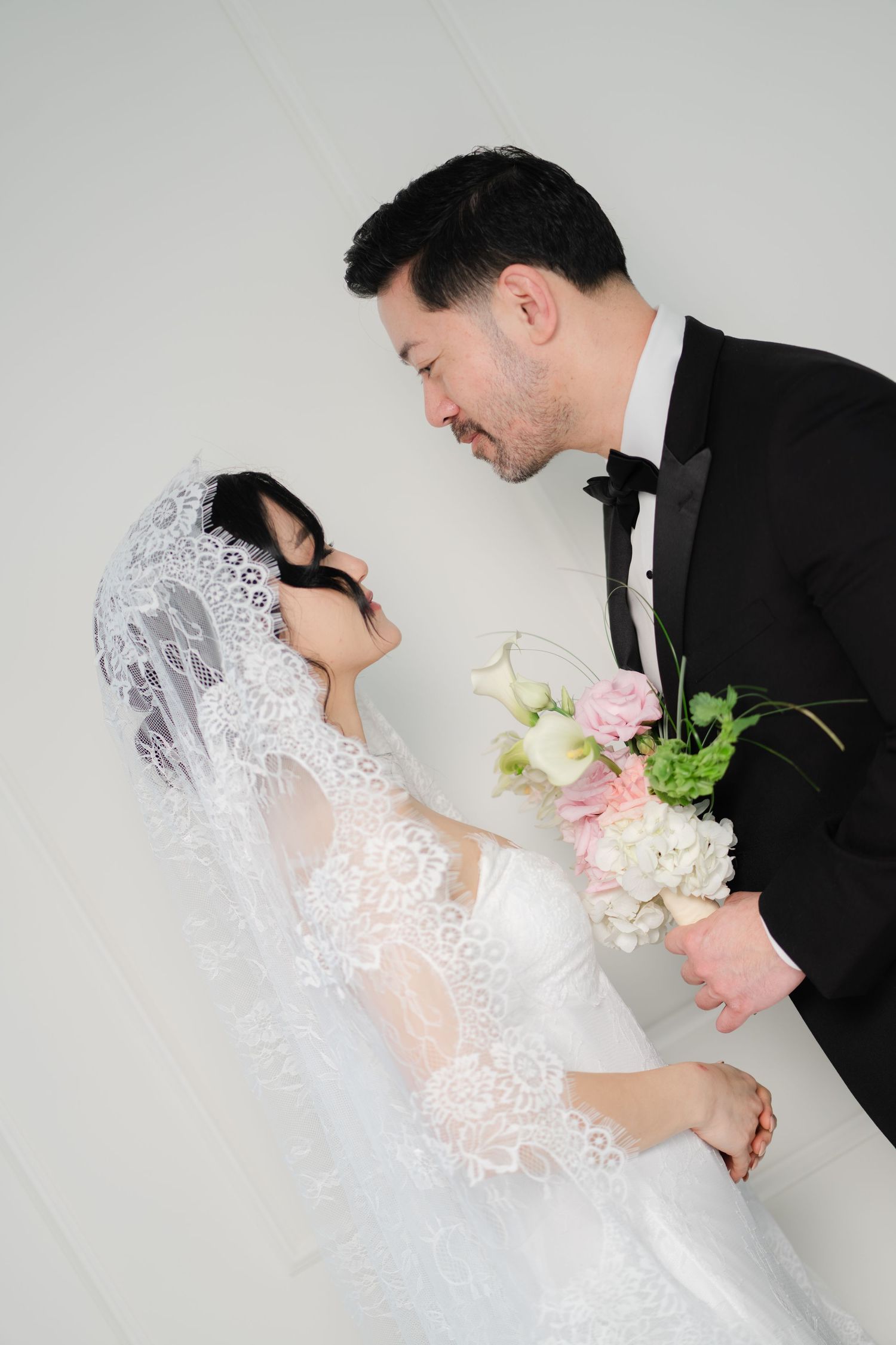 Asian bride in lace veil and groom in tuxedo sharing romantic moment holding pink and white wedding bouquet.