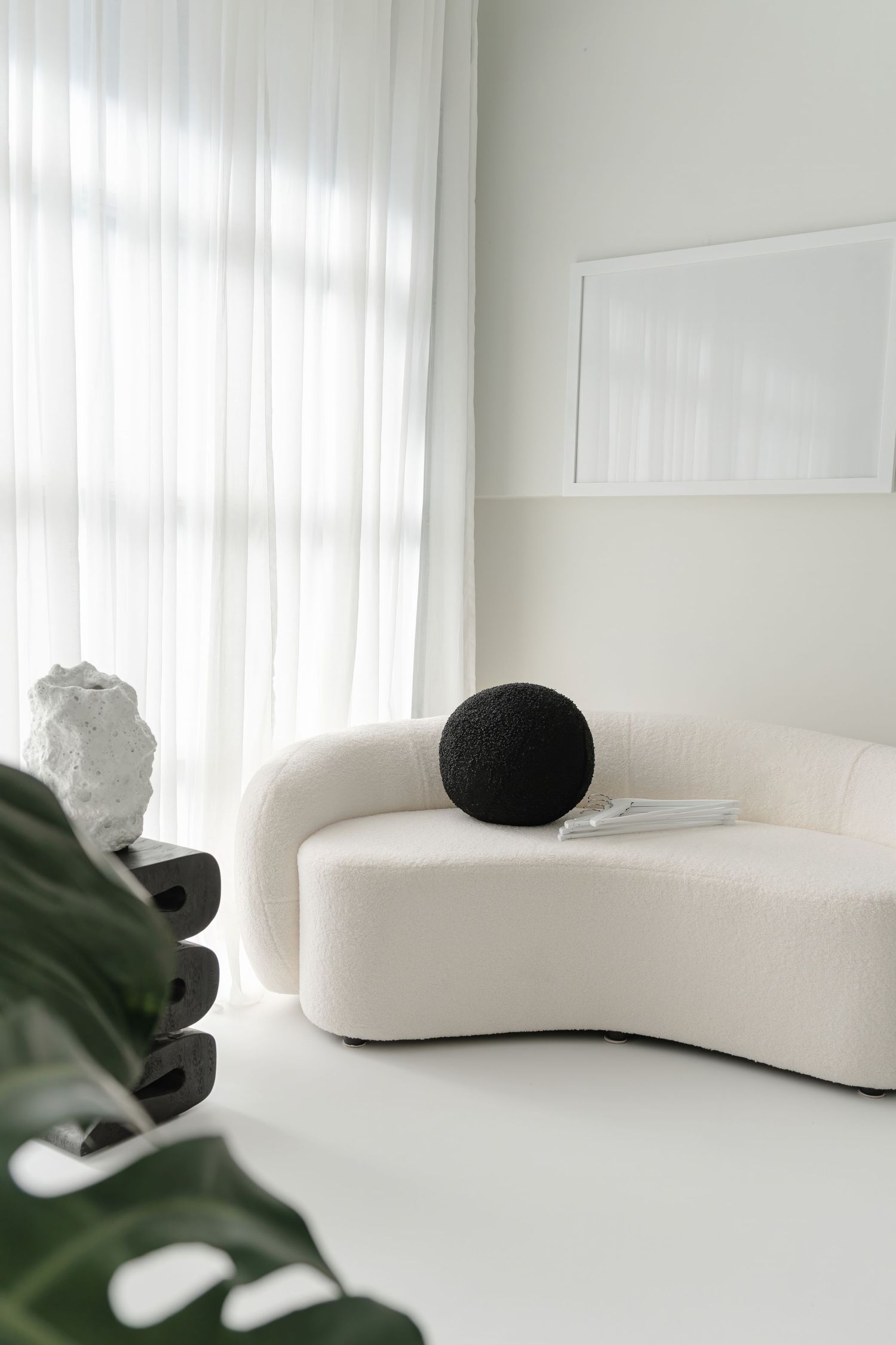 Curved white sofa with black sphere accent and monstera plant leaf in foreground.