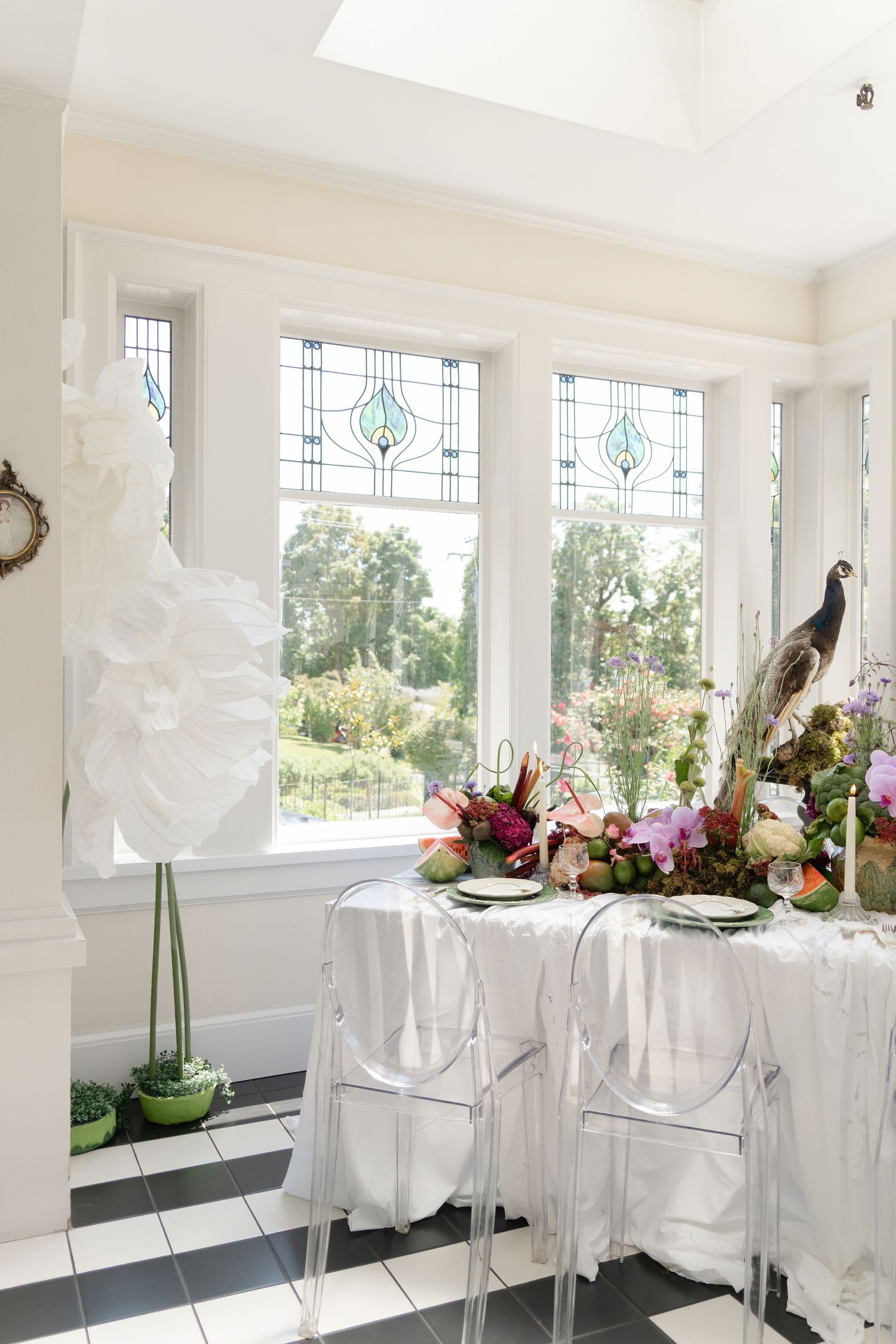Elegant wedding reception table setup with white chairs and floral centerpieces in a bright sunlit room with decorative windows.